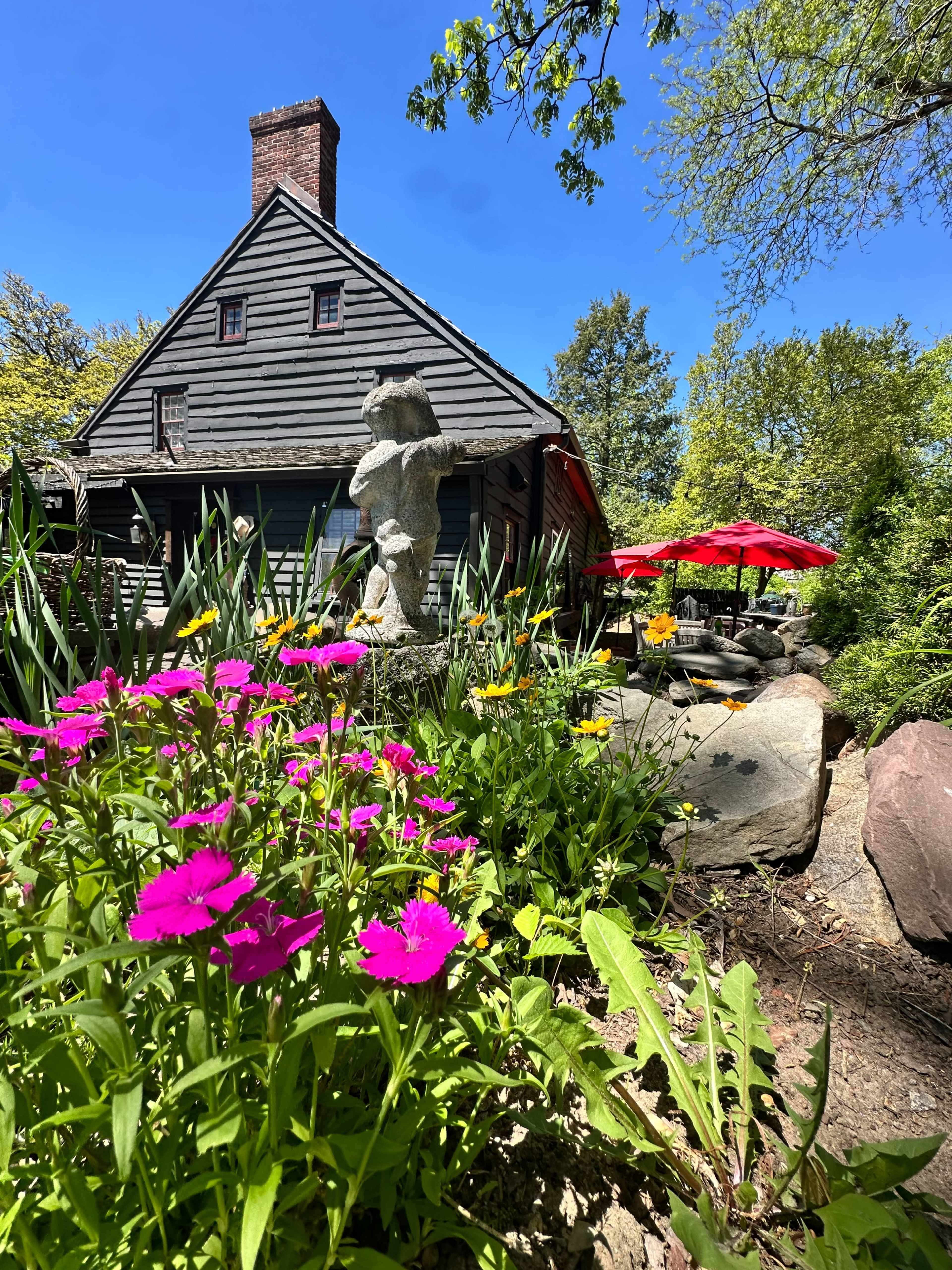 A rustic wooden house is situated behind a garden filled with colorful flowers and a stone sculpture, with a shaded patio area featuring red umbrellas.