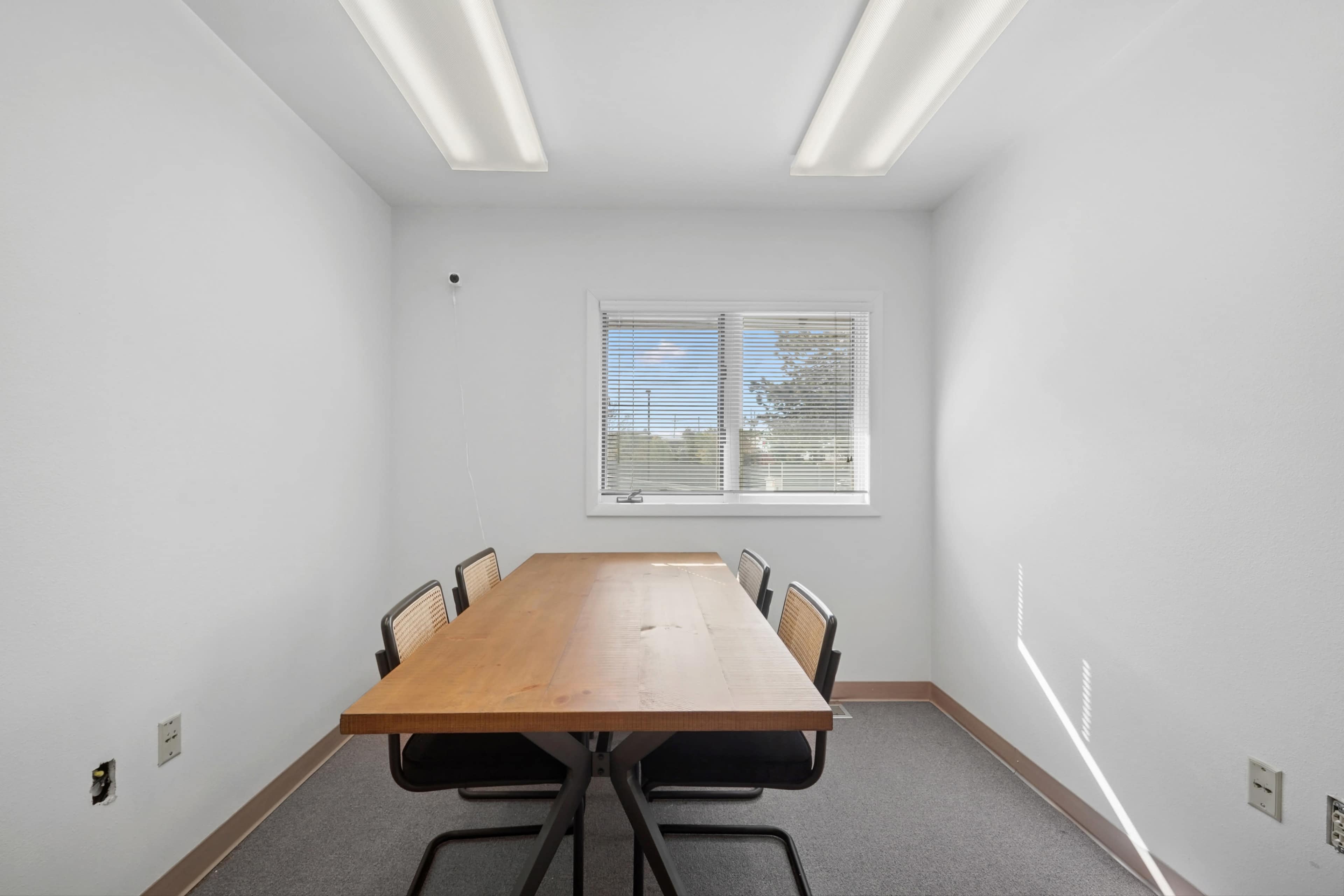 A small, brightly lit meeting room features a wooden table with four black chairs and a window providing natural light.