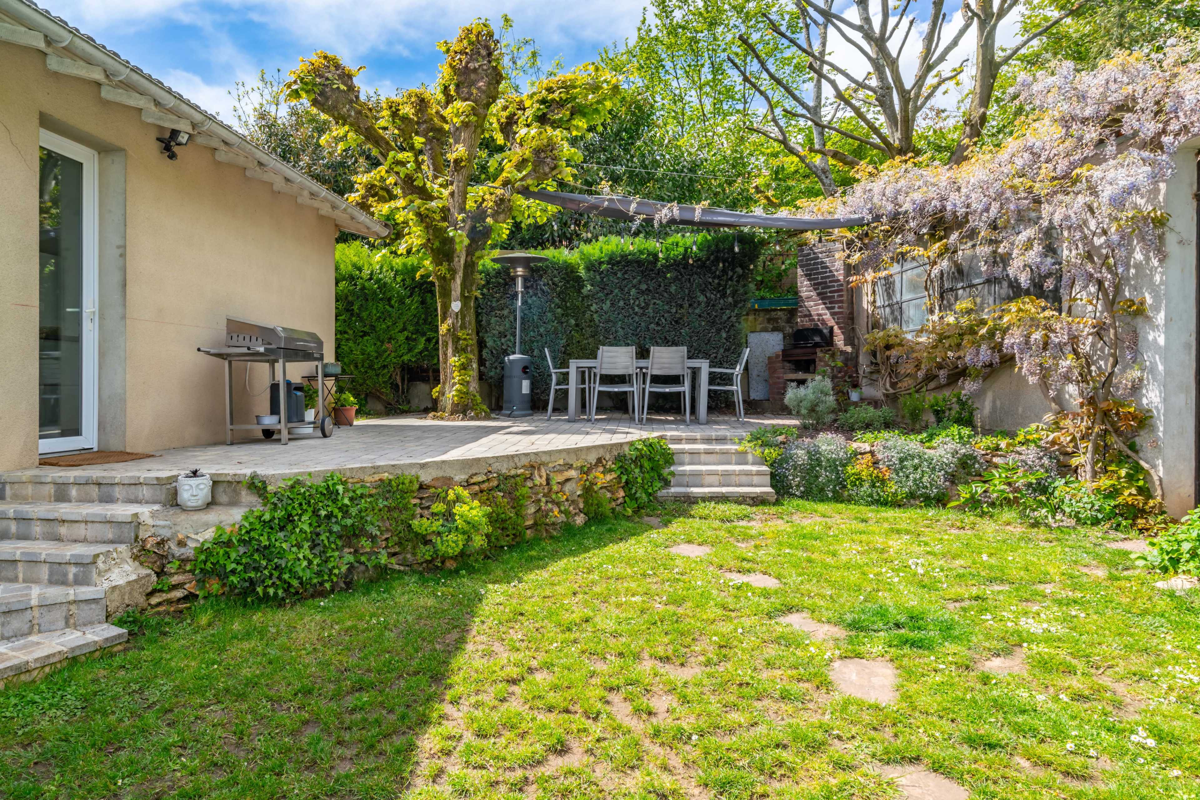 A wooden deck with a grill overlooks a landscaped garden featuring stone steps, seating area, and flowering plants.