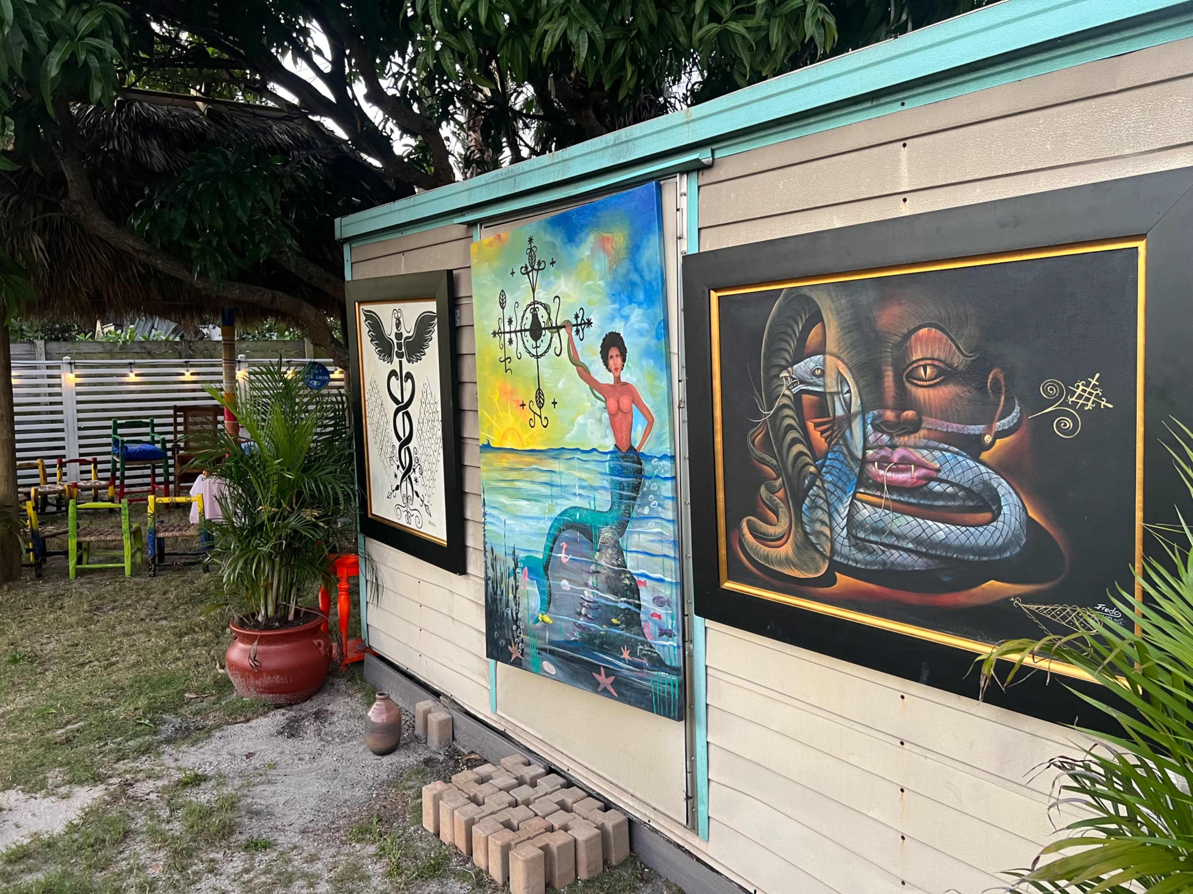 A wooden wall adorned with three murals, featuring vibrant designs including a mermaid, a snake, and symbolic elements, set in a grassy outdoor area.