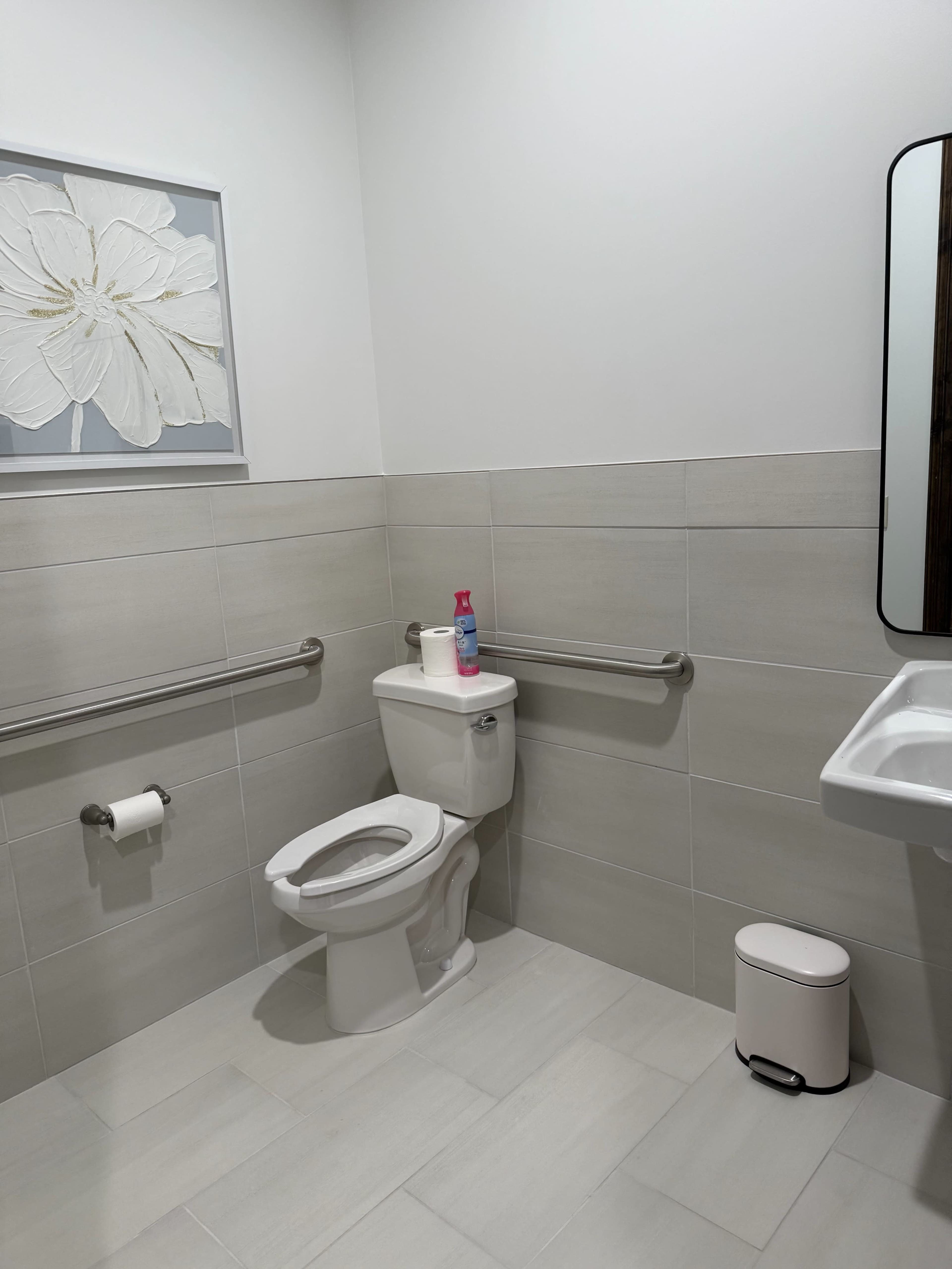 The image shows a clean, modern bathroom with a toilet, a sink, and a wall-mounted mirror, complemented by light-colored tiles and a flower-themed wall decoration.
