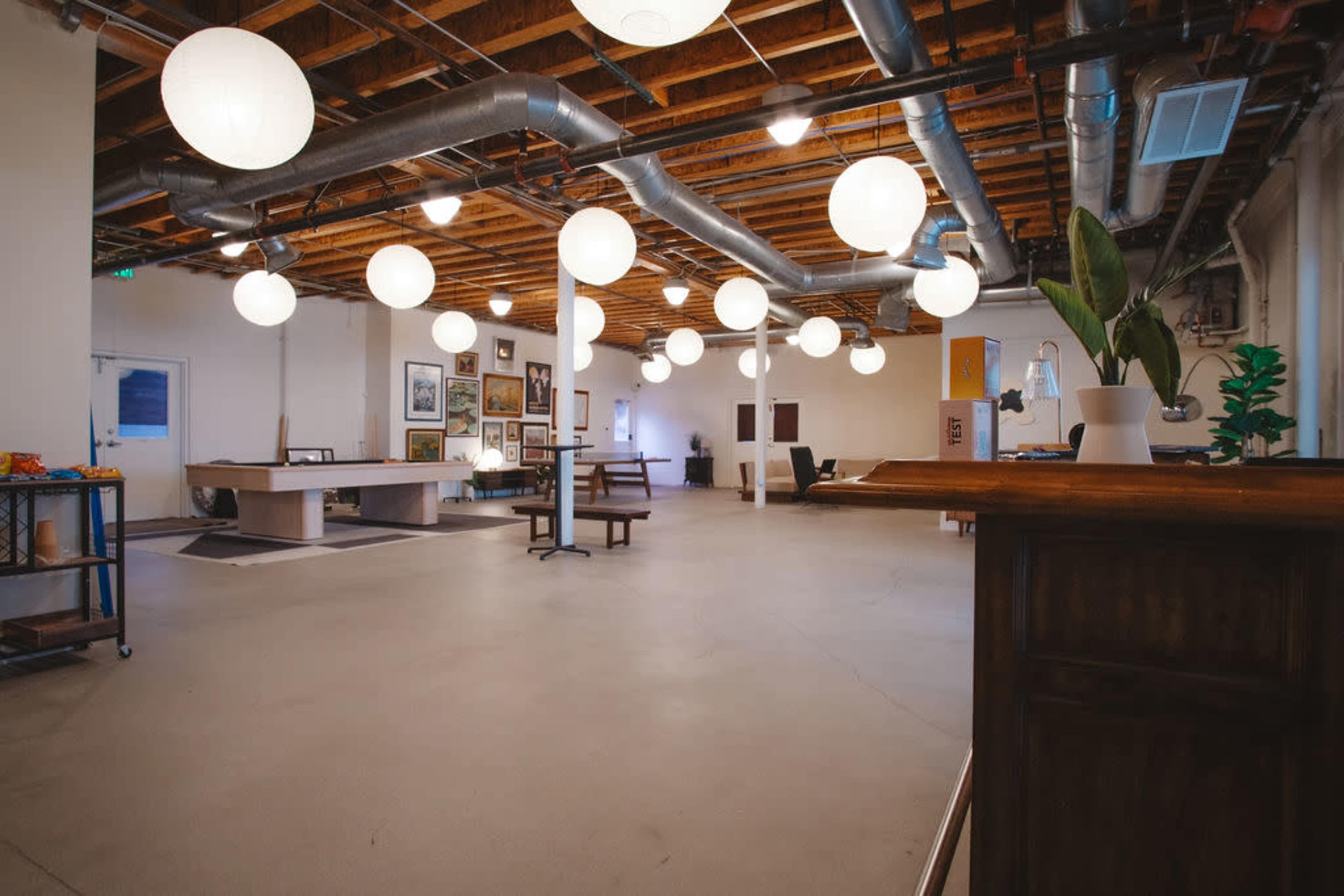 The image shows a spacious interior of a room featuring a high ceiling with industrial pipes, multiple round light fixtures, and a mixture of furniture, including tables and seating areas.