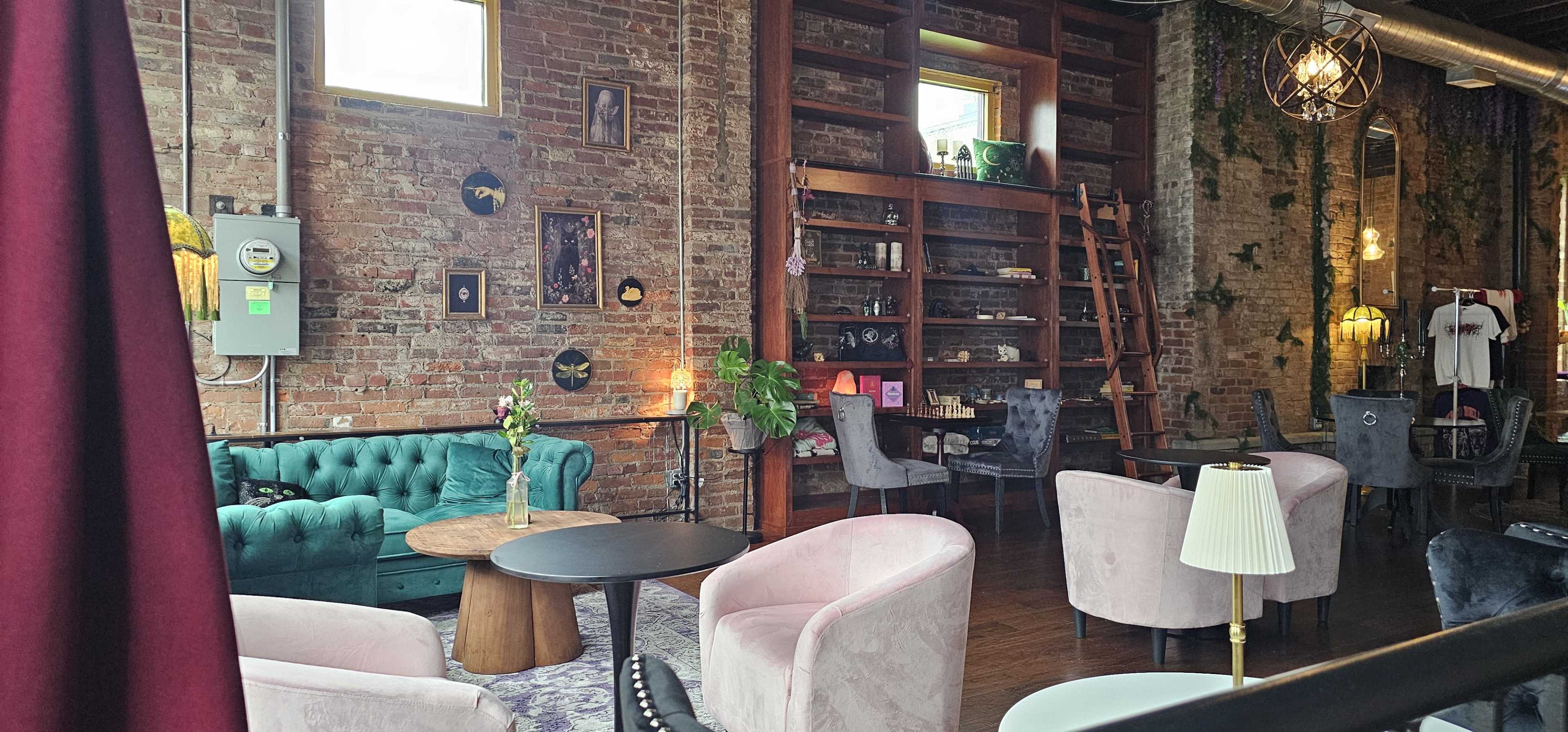 The image shows a cozy café with exposed brick walls, green plants, and a mix of seating options including plush chairs and a tufted sofa.
