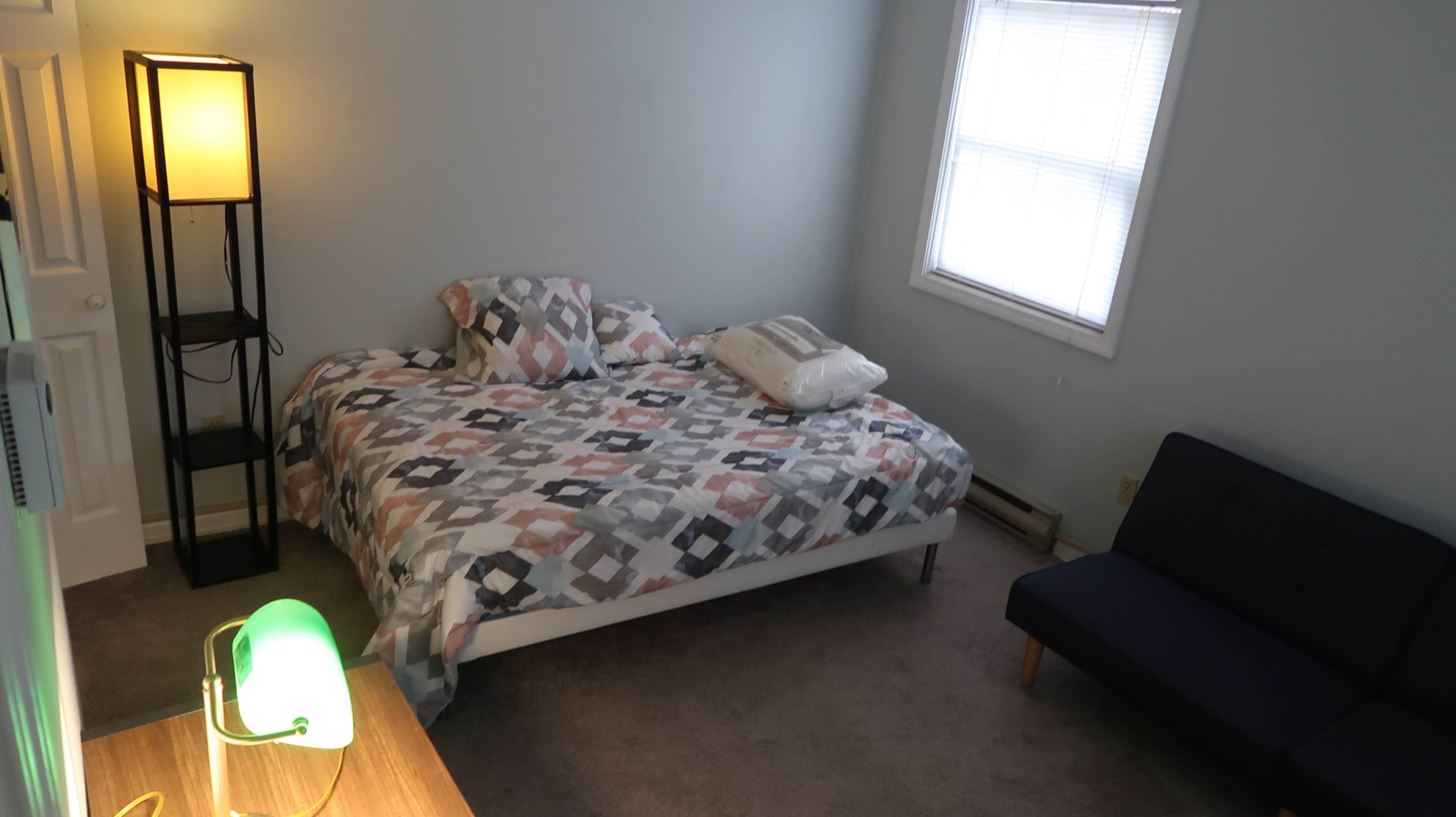 The image shows a bedroom with a bed covered in a patterned quilt, a lamp, a small blue couch, and a window allowing natural light.