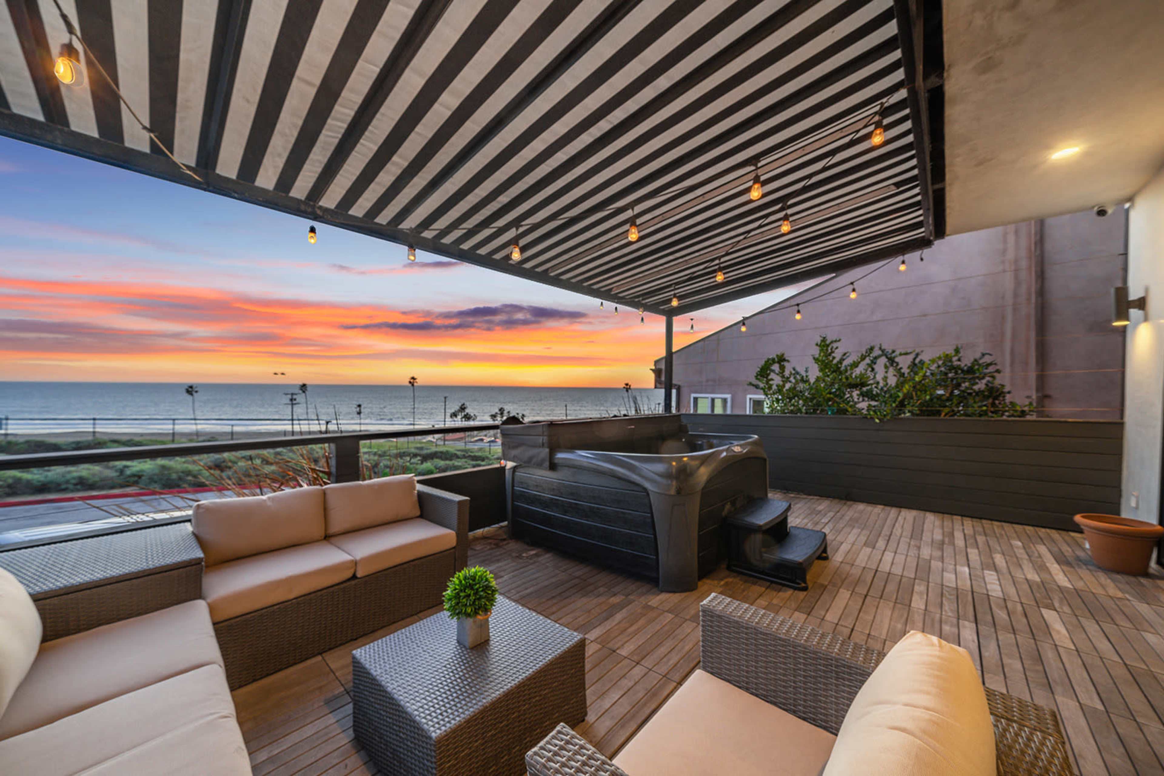 A patio with striped awnings features a hot tub and comfortable seating, overlooking a sunset view of the ocean.