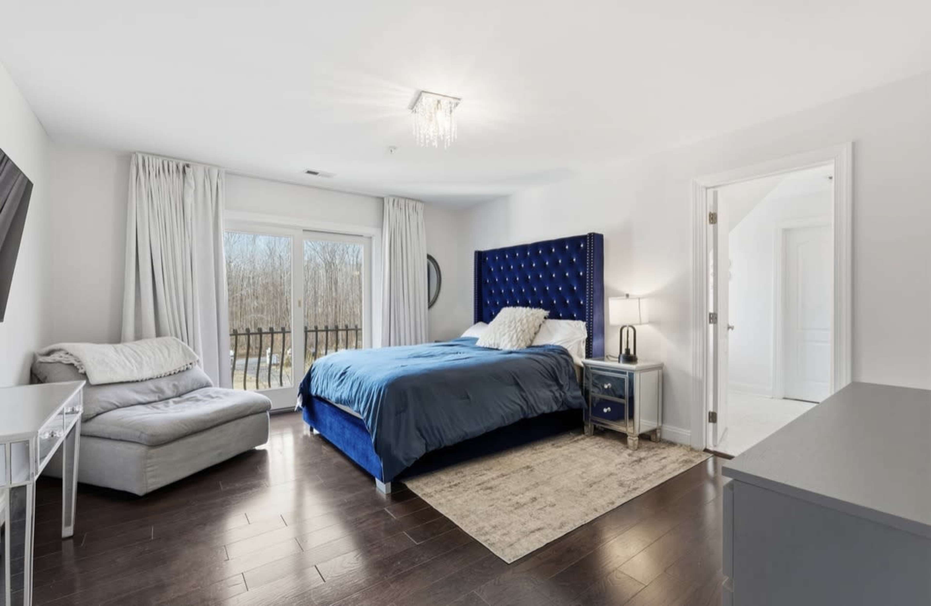 The image shows a modern bedroom with a blue upholstered bed, a plush armchair, and large windows overlooking a wooded area.