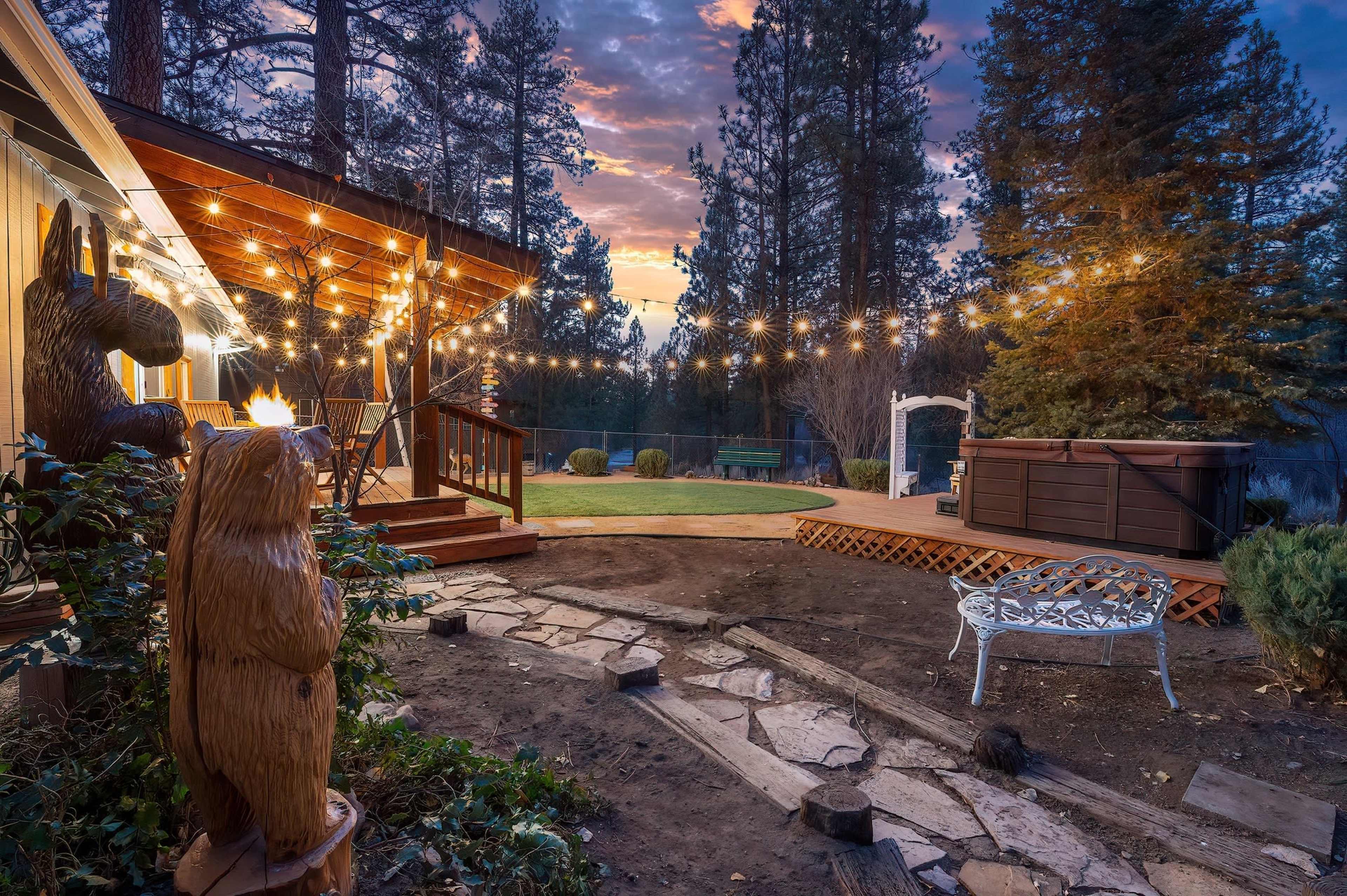 A wooden deck with string lights overlooks a landscaped yard featuring a hot tub and a bear sculpture amidst tall pine trees at sunset.