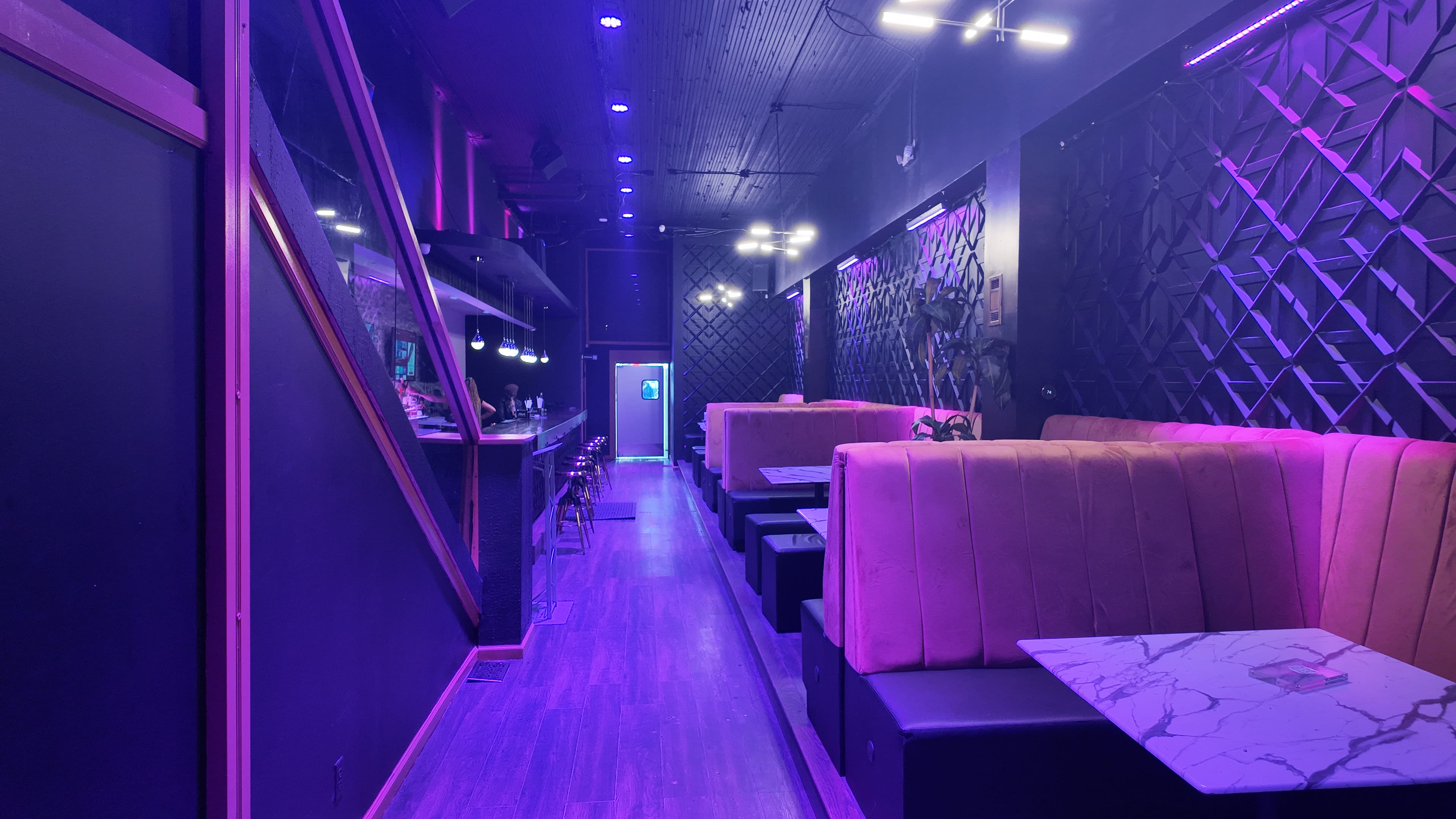 The image shows a modern interior of a dimly lit lounge with purple lighting, featuring plush seating and a bar area along one side.