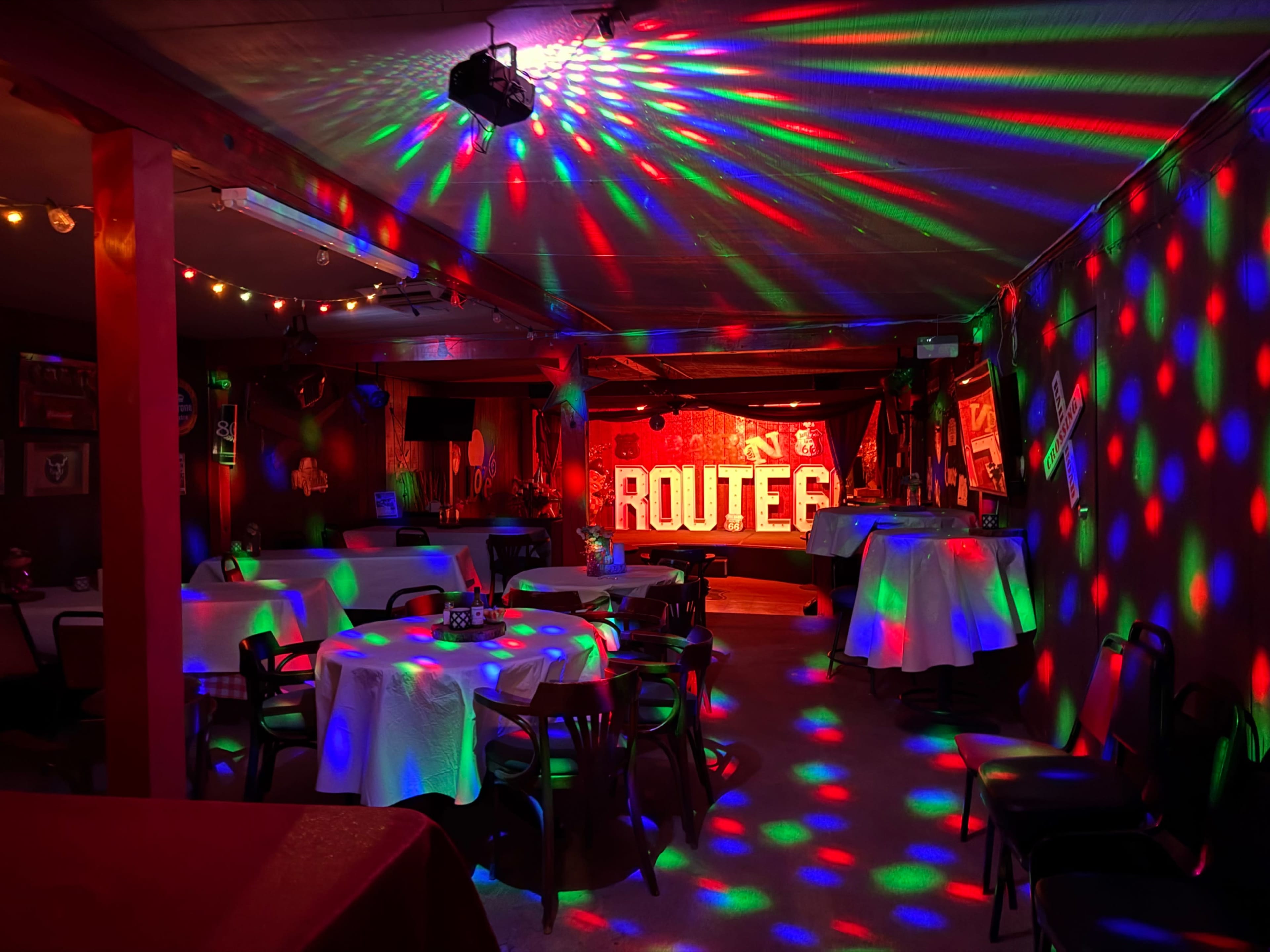 The image shows a dimly lit room decorated with colorful lights casting patterns on tables and walls, featuring a backdrop that reads "ROUTE 66."