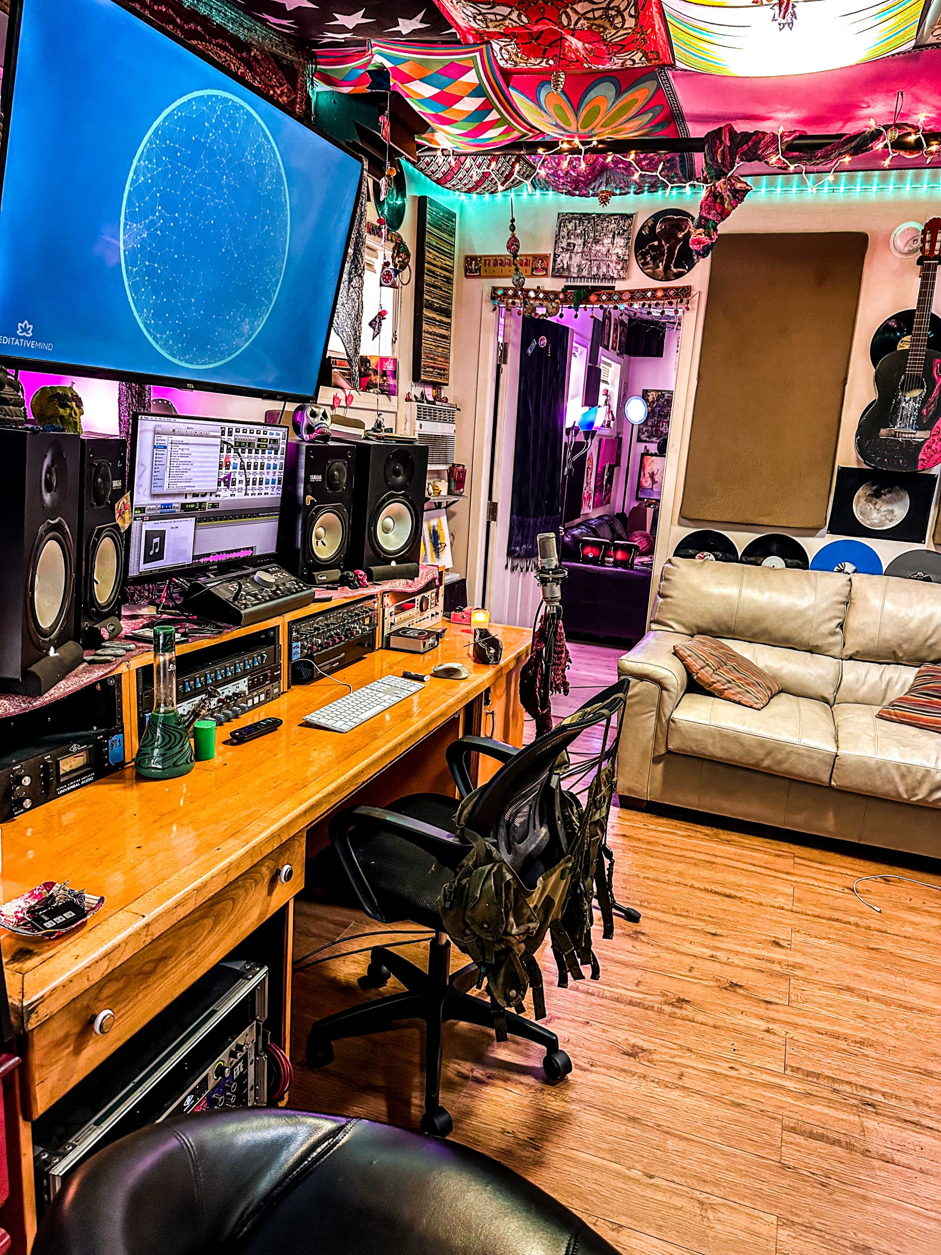 The image shows a colorful music studio with a wooden desk, various audio equipment, large speakers, and a couch, all under a decorated ceiling.