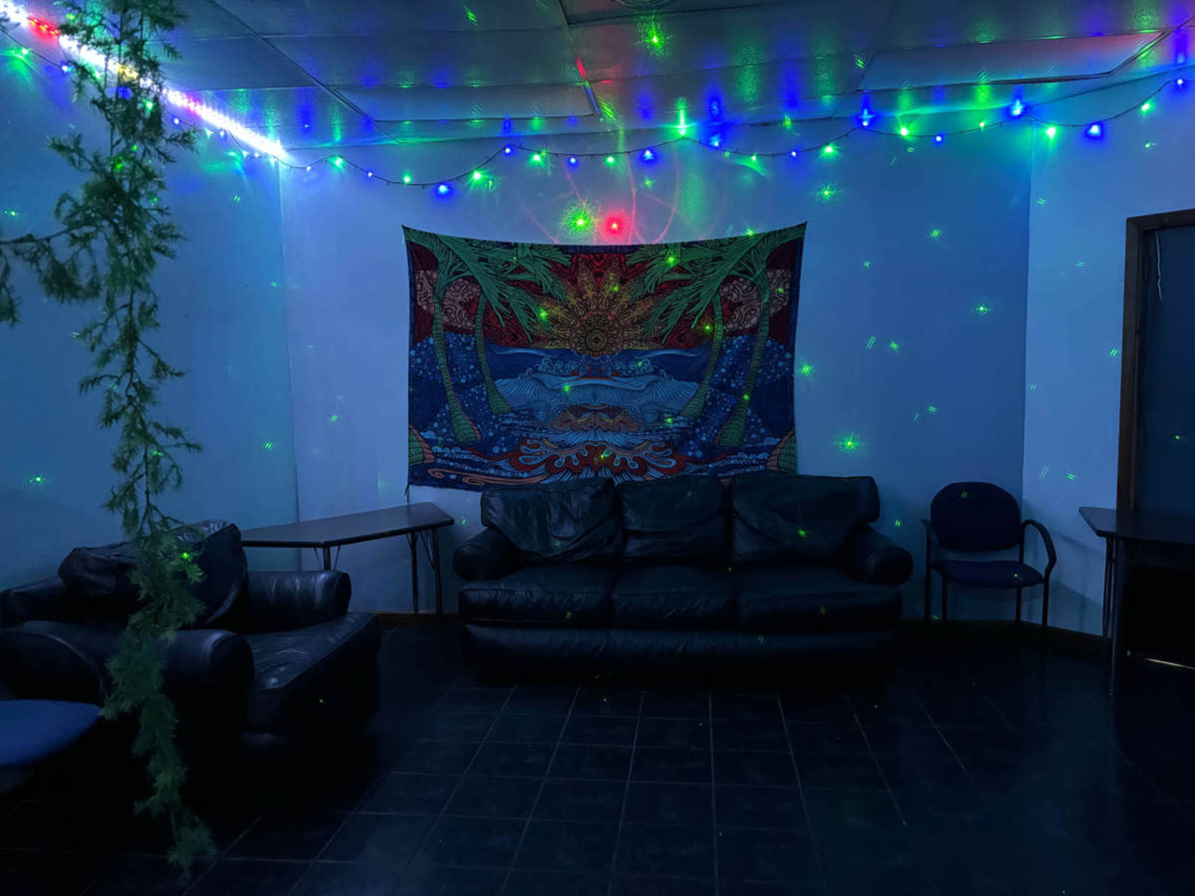 A dimly lit room features a black couch, blue chairs, and a colorful tapestry hanging on the wall, illuminated by multicolored string lights and a laser projector.