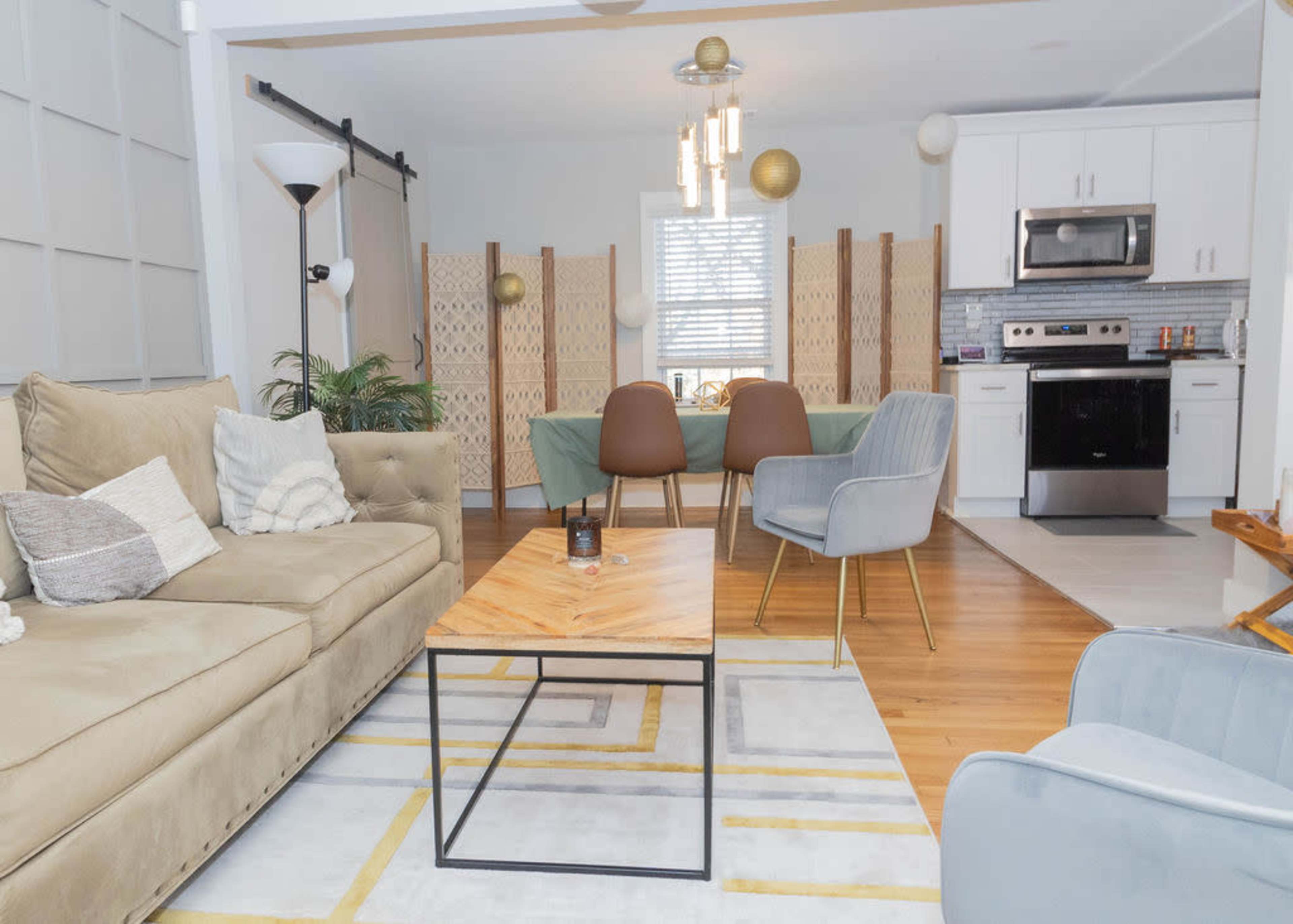 The image shows a modern living space featuring a beige couch, a wooden coffee table, and a dining area with a green tablecloth, surrounded by decorative screens and kitchen appliances.
