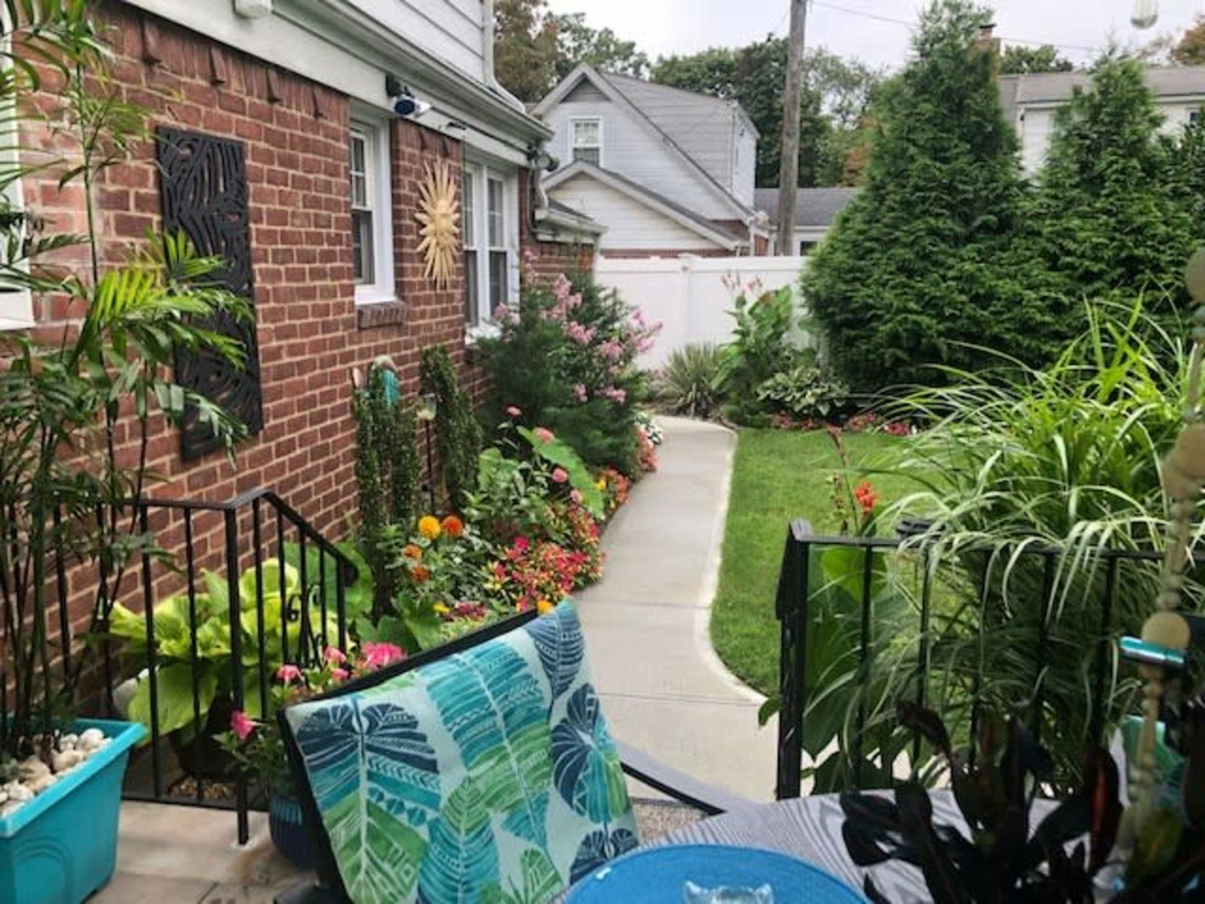The image shows a residential exterior with a pathway leading through a garden filled with colorful flowers, bordered by greenery and a brick wall.