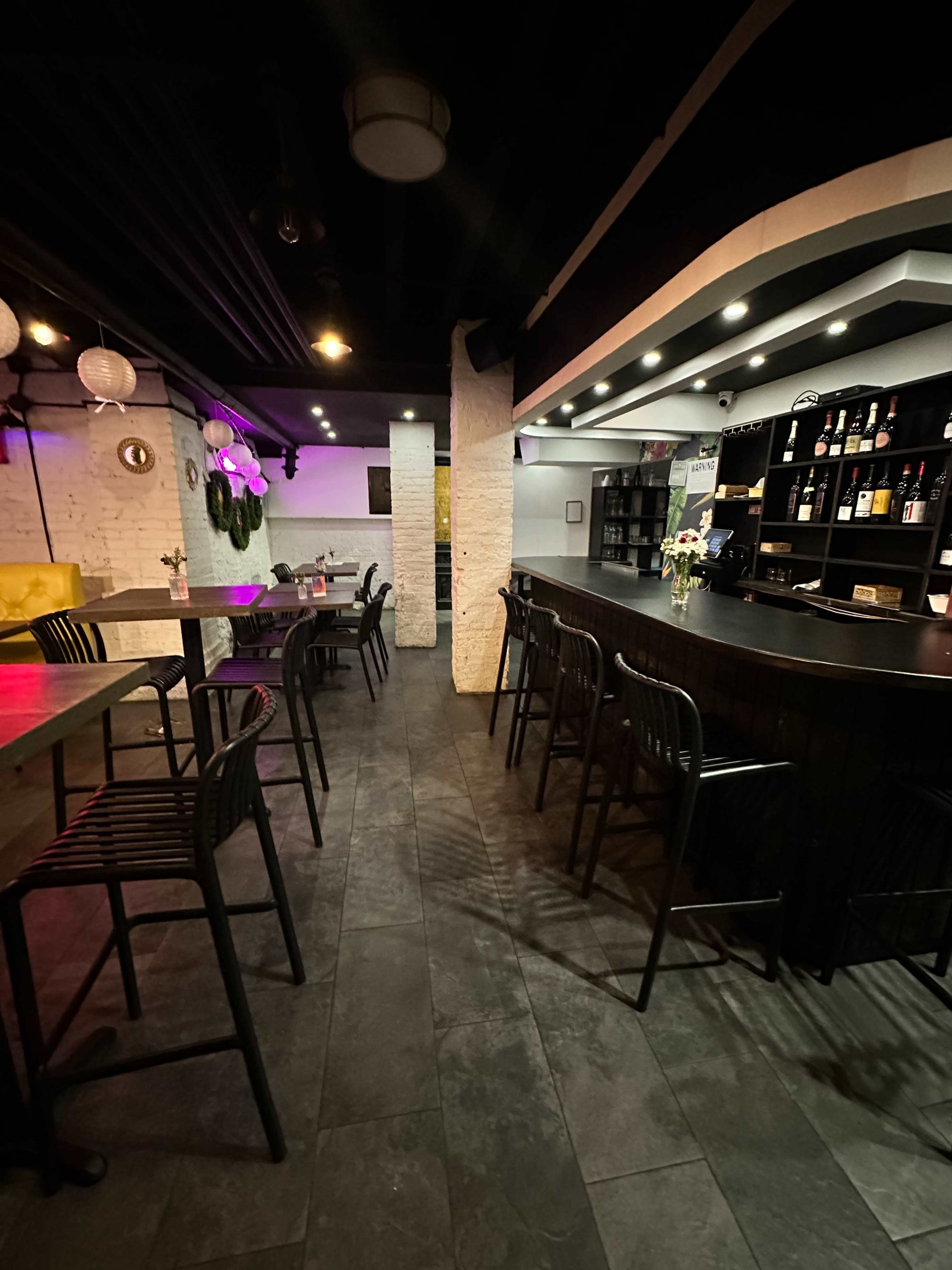A dimly lit bar area with black furniture, a counter displaying various bottles, and tables arranged for seating.