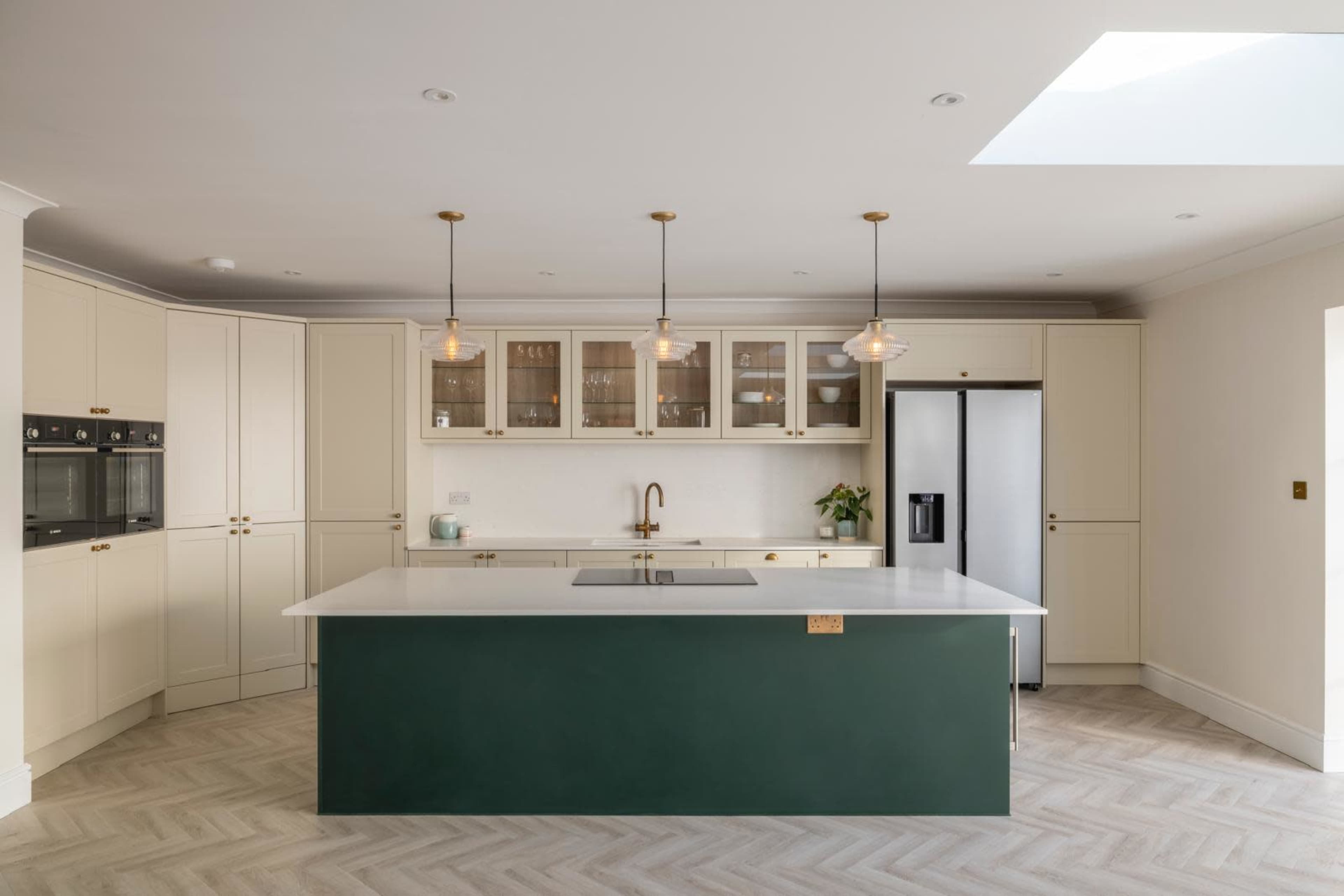 The image shows a modern kitchen with light-colored cabinets, a green kitchen island, and pendant lighting, featuring a skylight above.