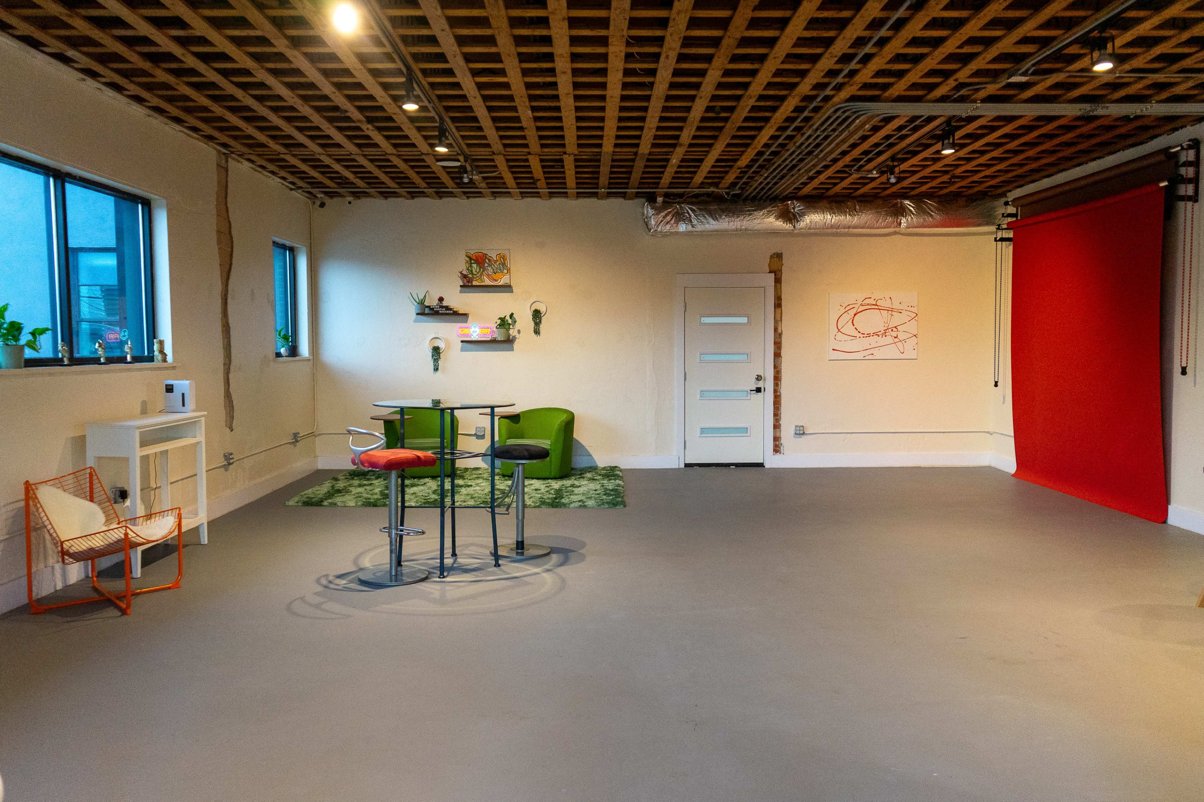 The image shows a spacious, minimalist room featuring a red backdrop, green chairs, a bar table with two stools, and a patterned rug, illuminated by natural light from nearby windows.