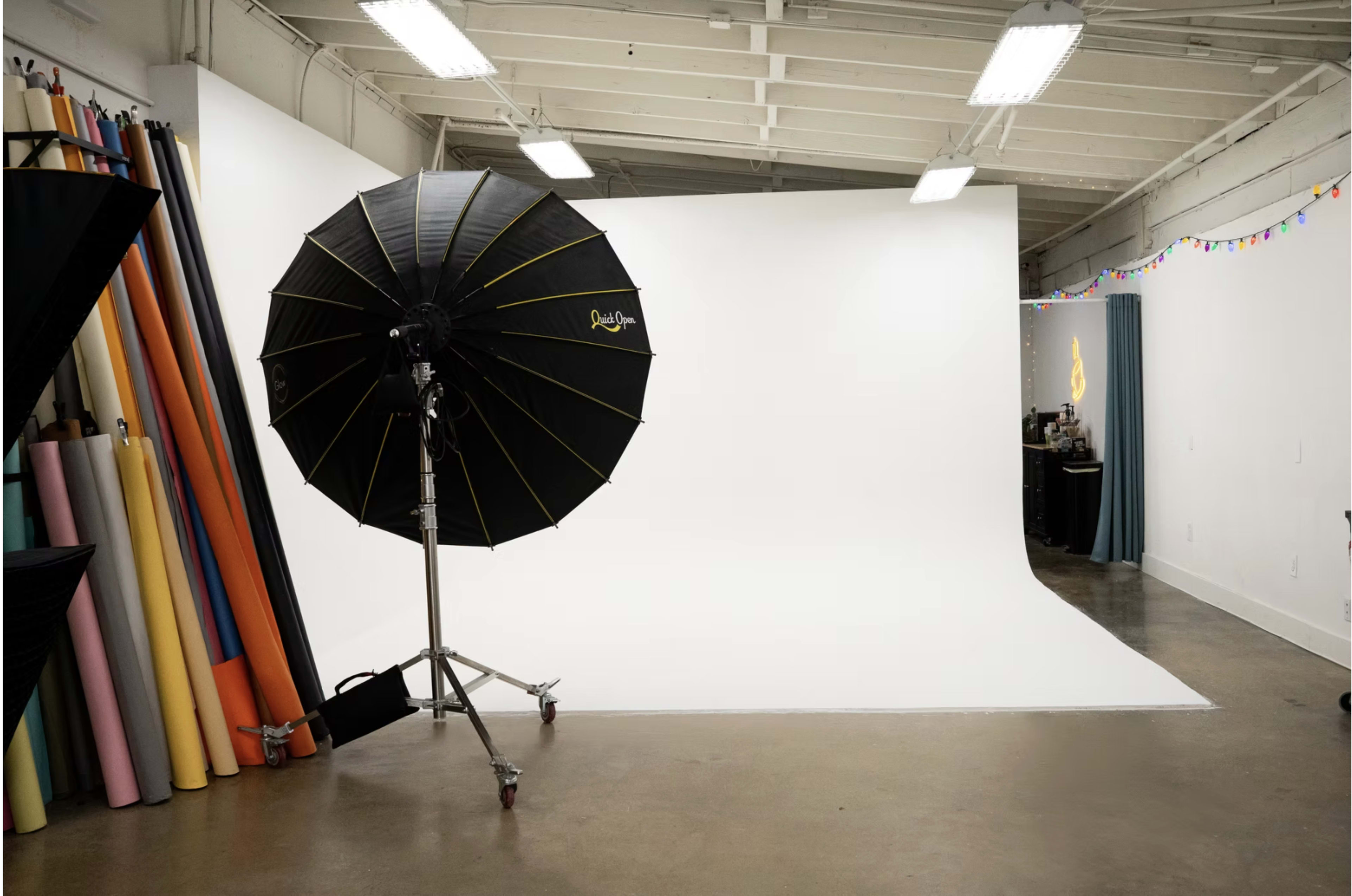 The image shows a photography studio with a white backdrop, a large black umbrella softbox, and rolls of colored backdrop papers along one side.
