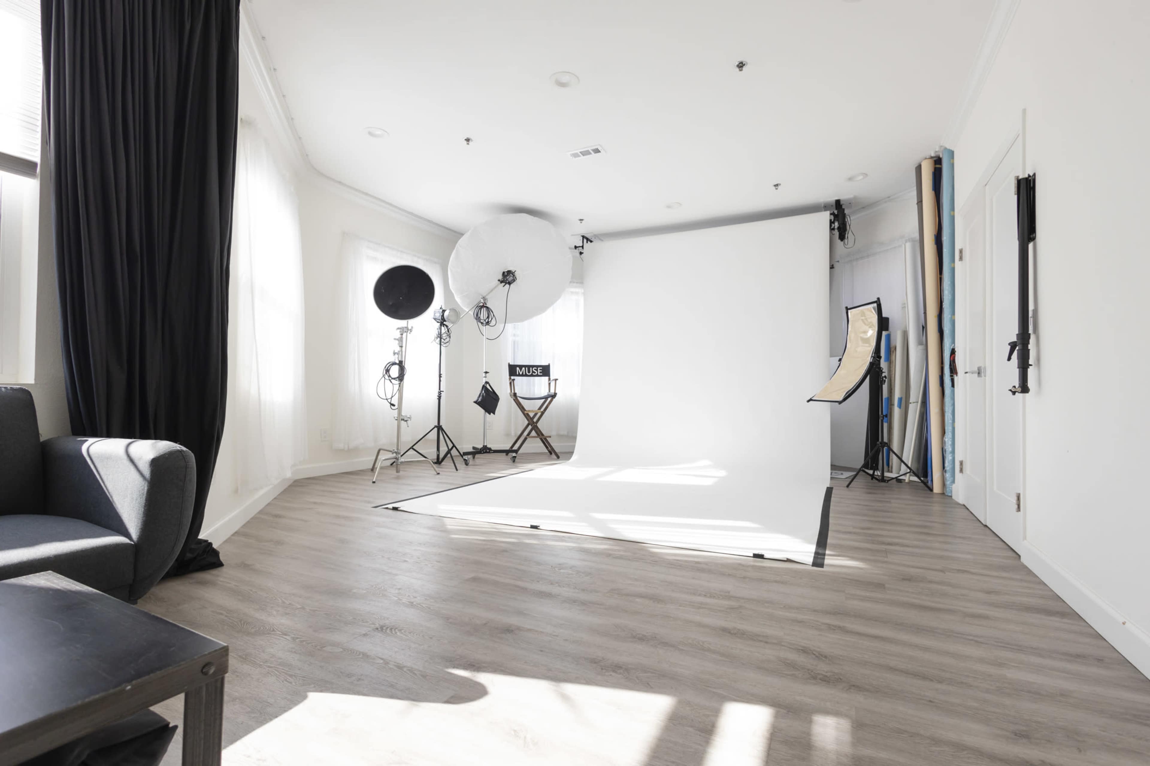 The image shows a photography studio with a large white backdrop, various lighting equipment, and a gray sofa in the corner.