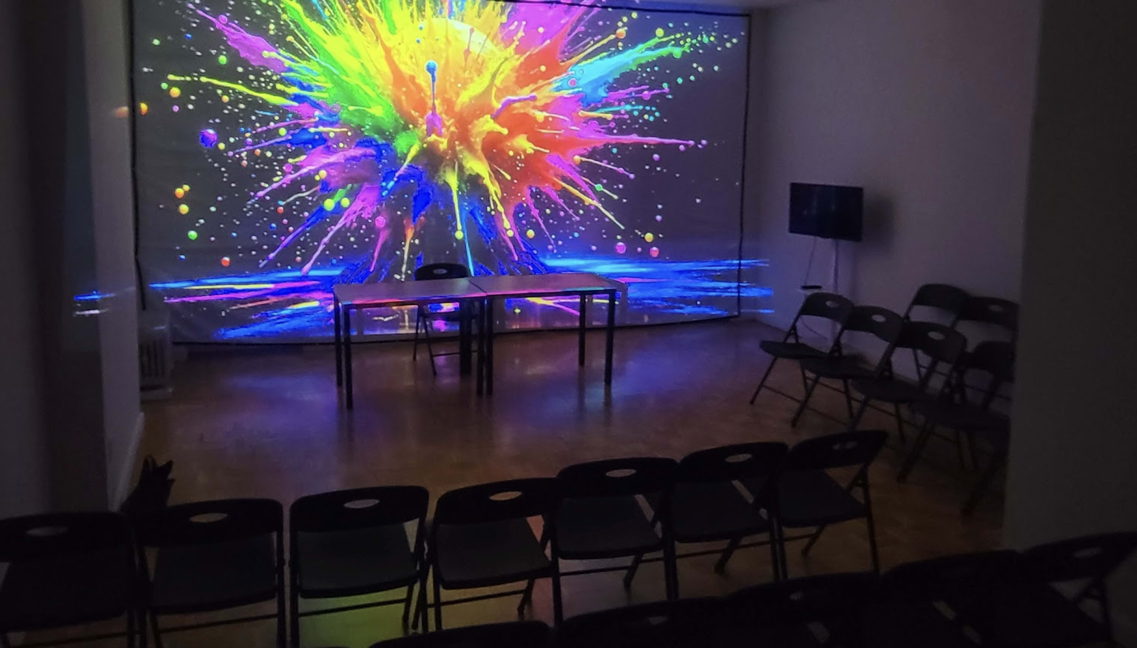 A brightly colored abstract splash projection covers the wall of a room with two tables in the center and folding chairs arranged around them.