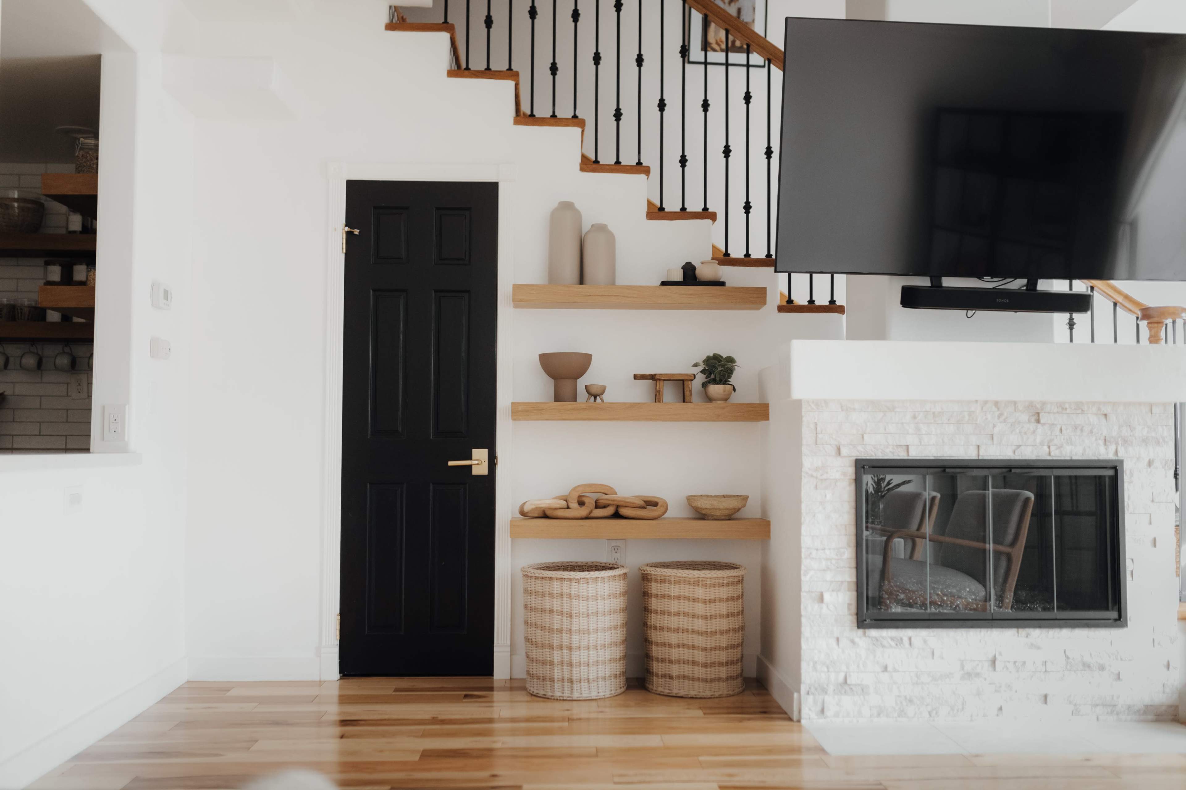 A modern living space featuring a staircase, a black door, shelves with decorative items, two woven baskets, and a fireplace with a television mounted above it.