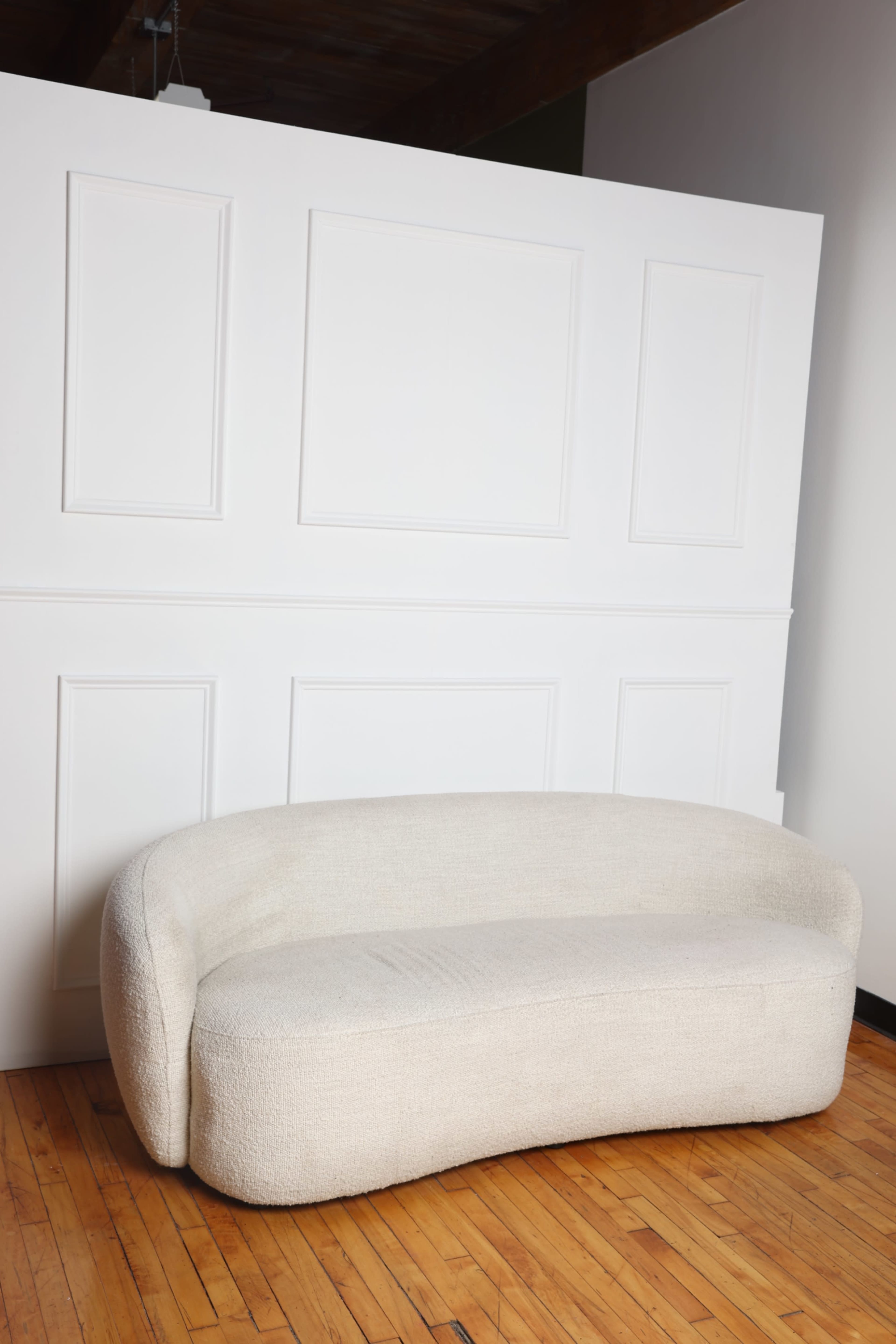 A curved, off-white sofa is positioned in front of a simple white paneled wall, placed on a wooden floor.