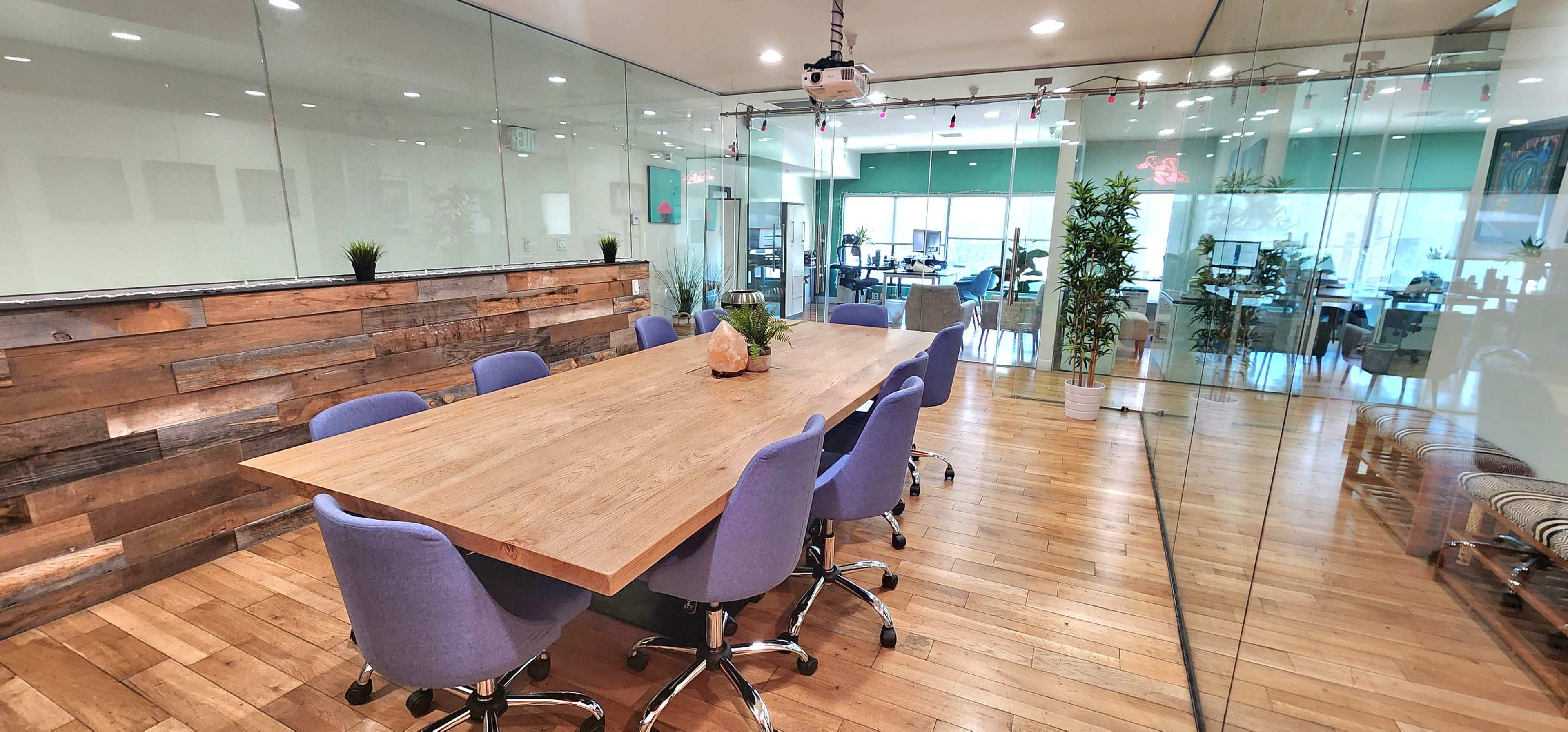 A large conference room features a wooden table surrounded by purple chairs, with glass walls that overlook an open office space.
