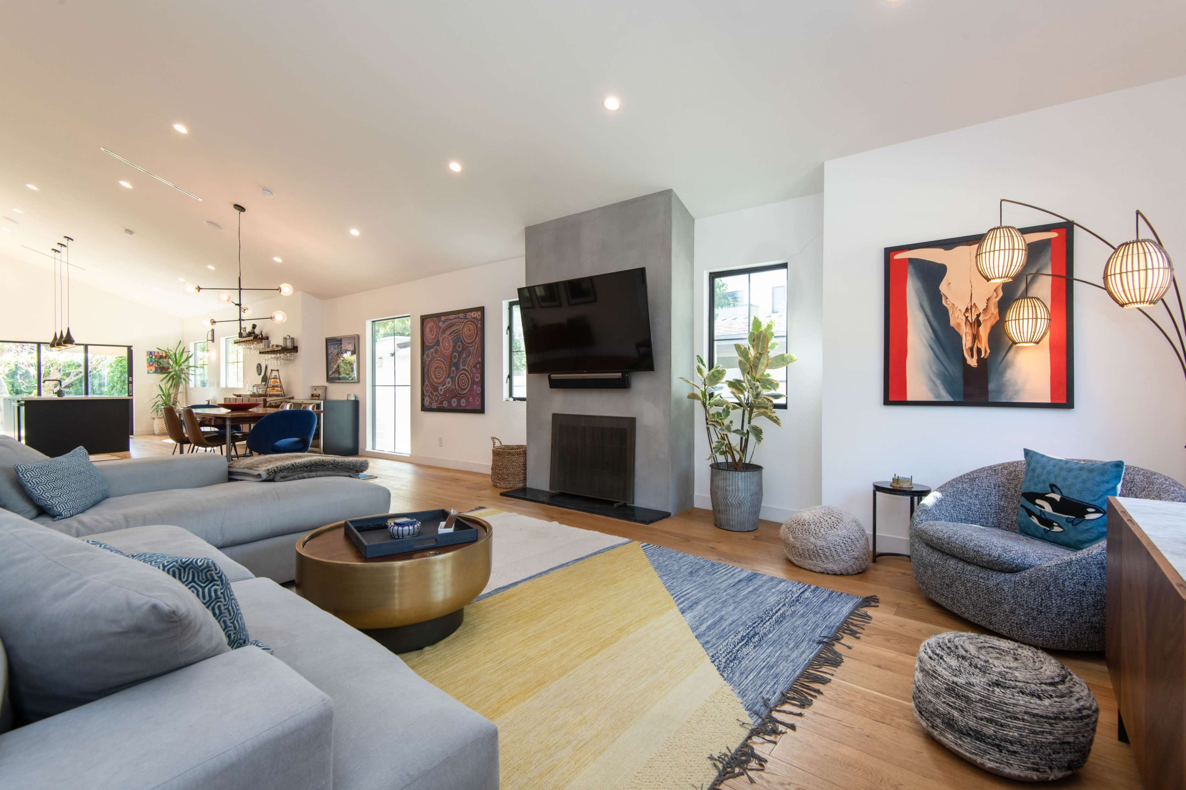 A modern living room features a sectional sofa, a large flat-screen TV mounted on a gray wall, a circular coffee table, and various decorative elements including artwork and potted plants.