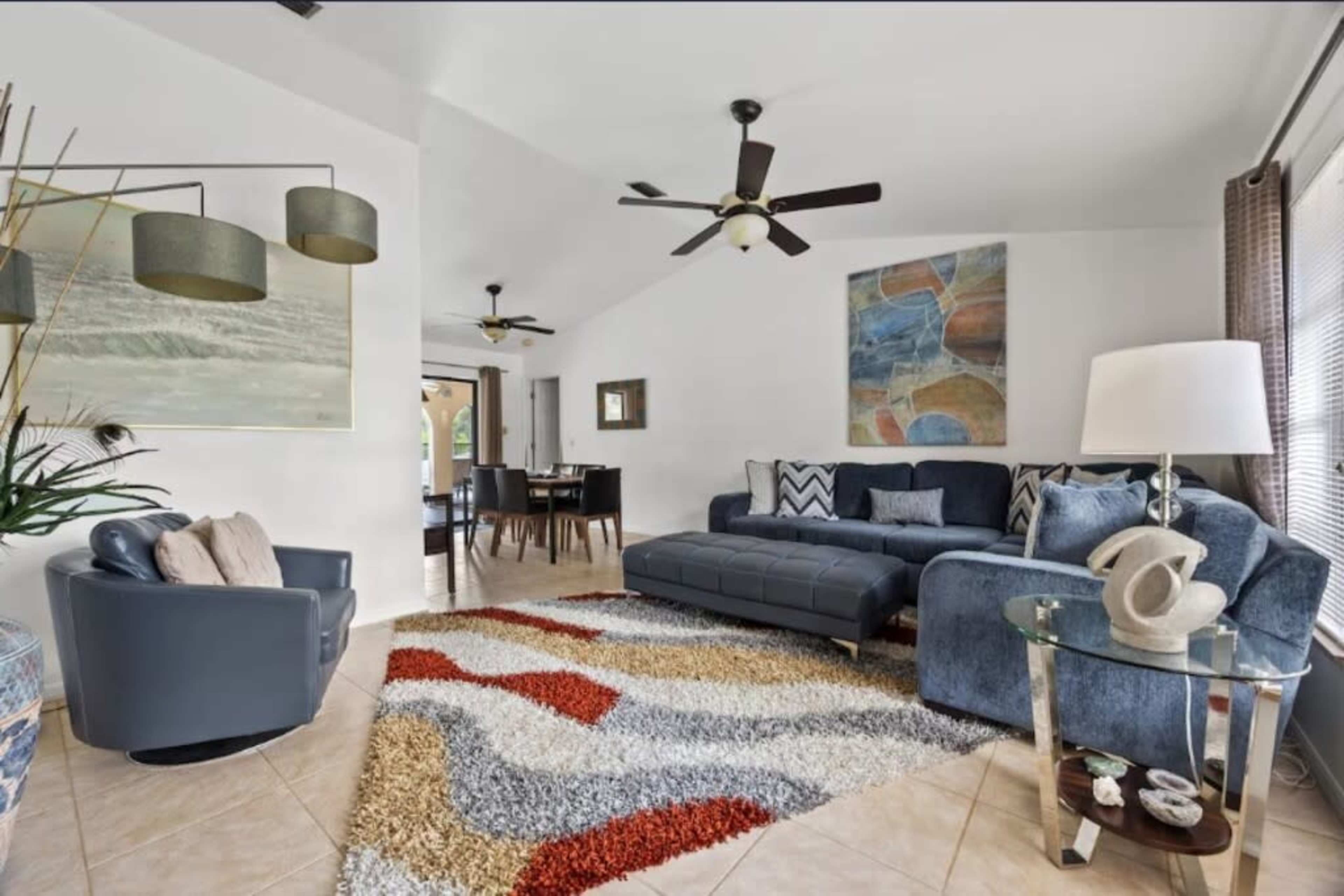 The image shows a modern living room with a blue sectional sofa, a round glass table, a patterned area rug, and an artwork on the wall, featuring a dining area in the background.