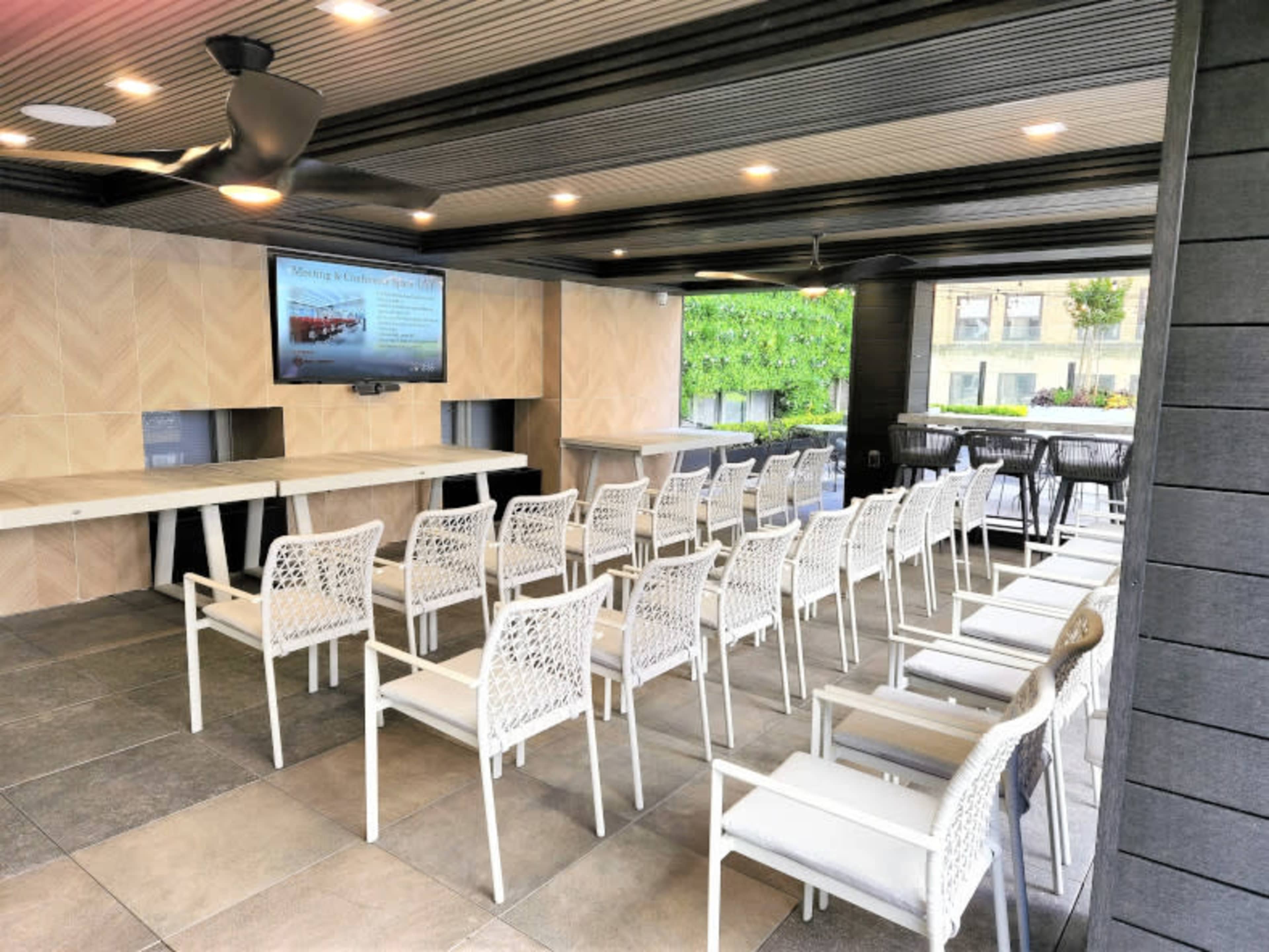 The image shows an indoor event space with rows of white chairs facing a wall-mounted television and a light-colored wood panel behind it.