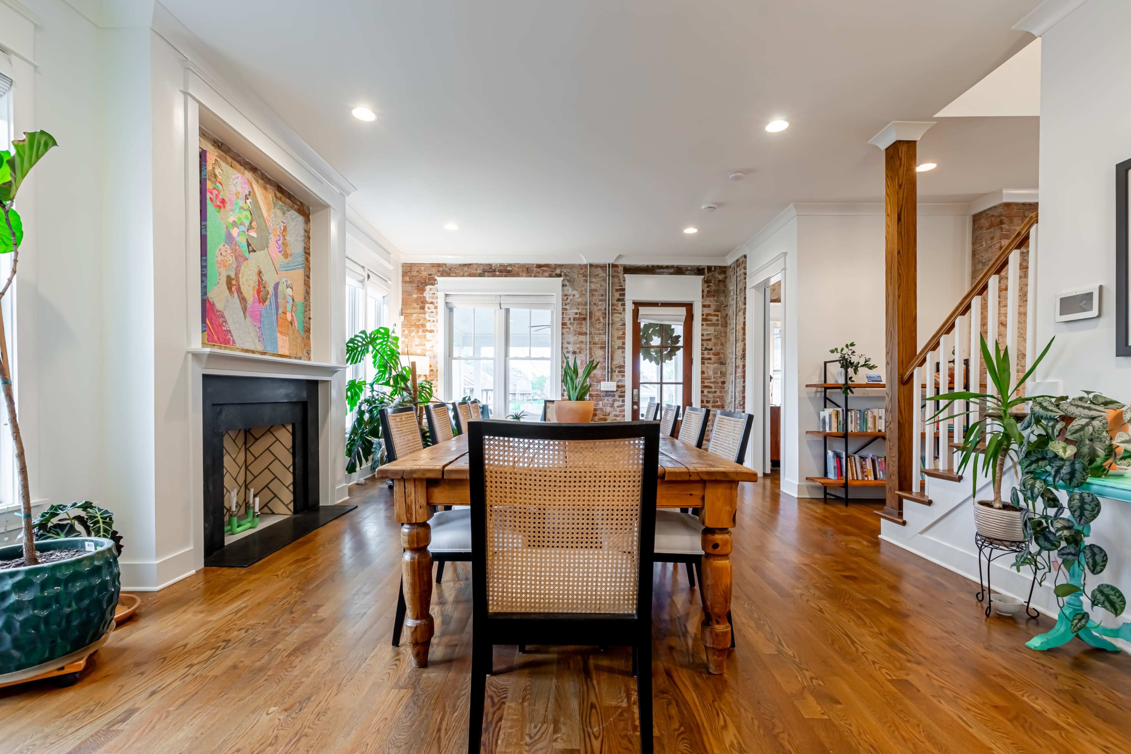 A spacious dining area features a long wooden table surrounded by chairs, with a fireplace and decorative plants in a bright, open layout.