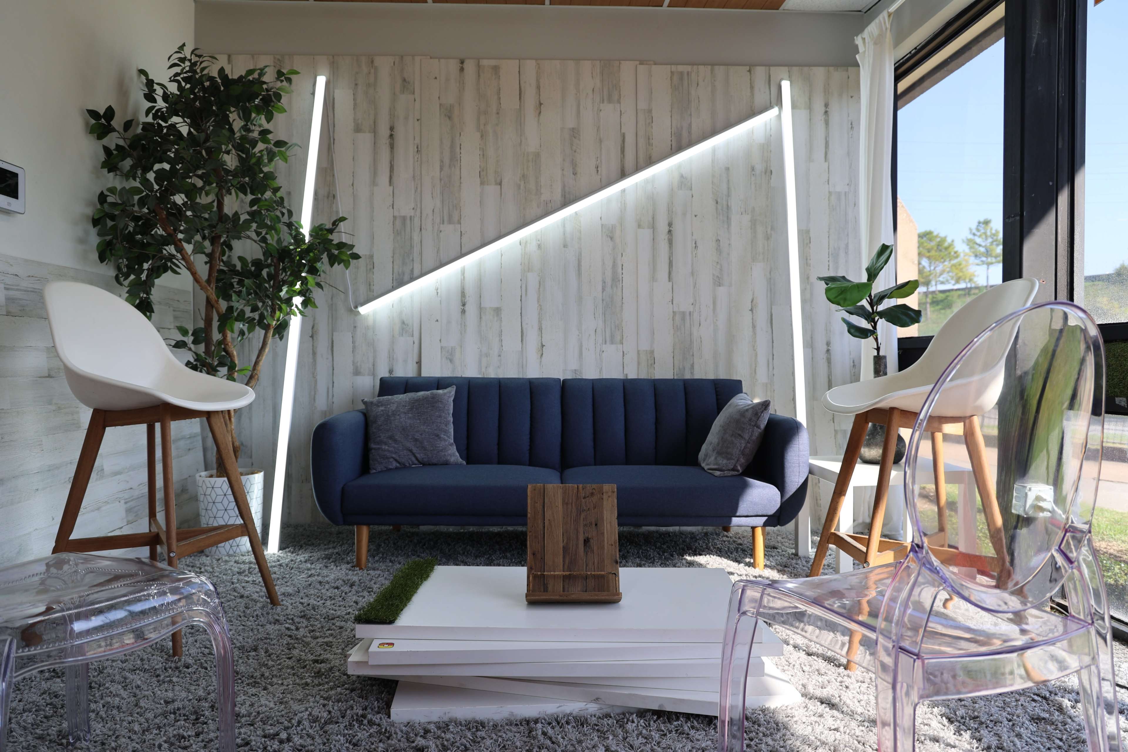 A modern living room features a blue sofa, clear chairs, and a wooden table, with plants and angled lighting against a textured wall.