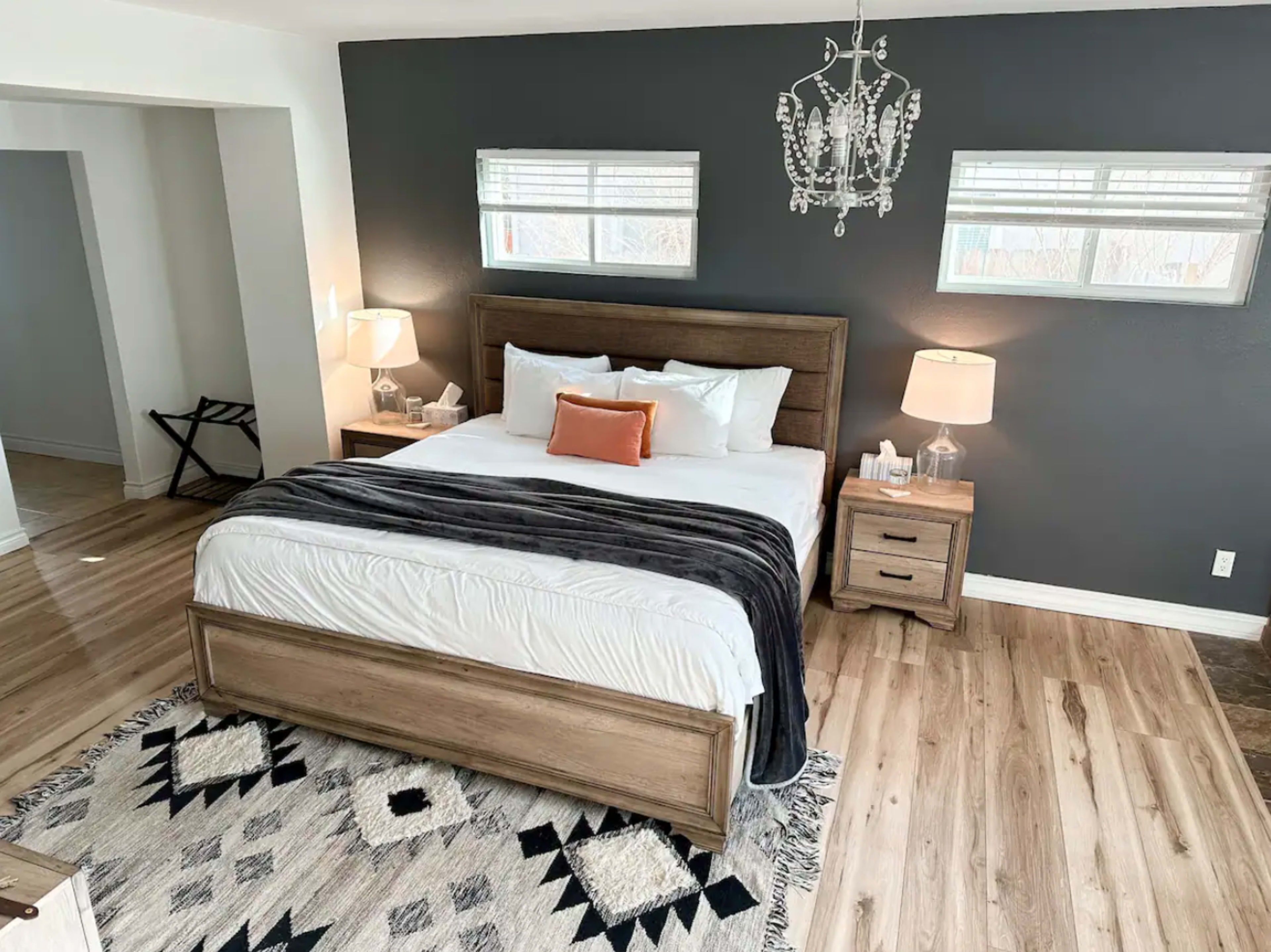 A modern bedroom with a large wooden bed, two bedside tables with lamps, and a chandelier, all set against a dark gray accent wall.