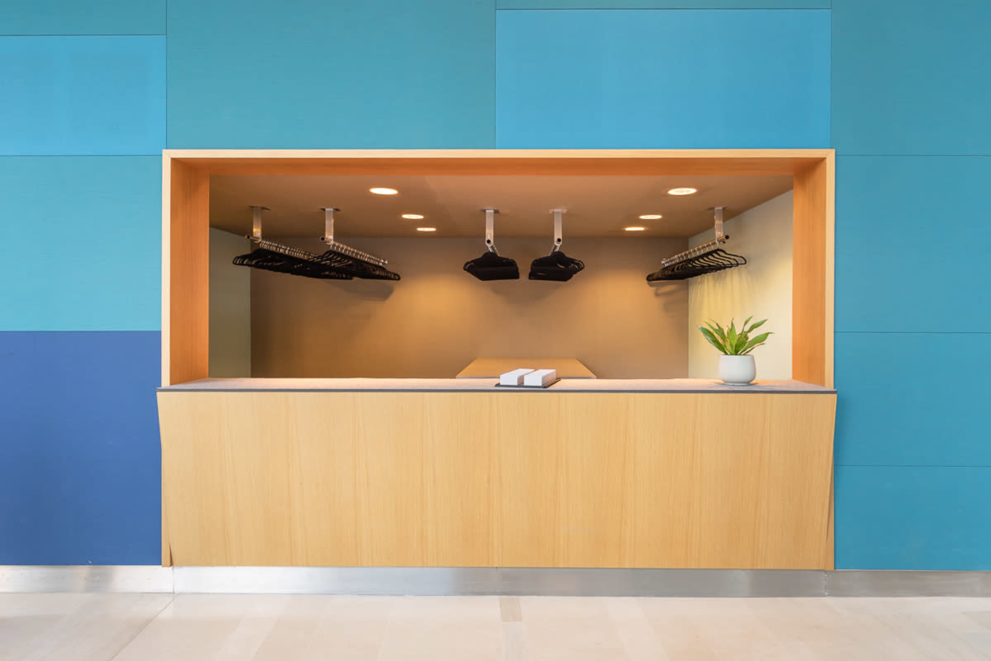 The image shows a modern reception area with a wooden counter, hanging coat racks, and a small potted plant in a minimalist design.