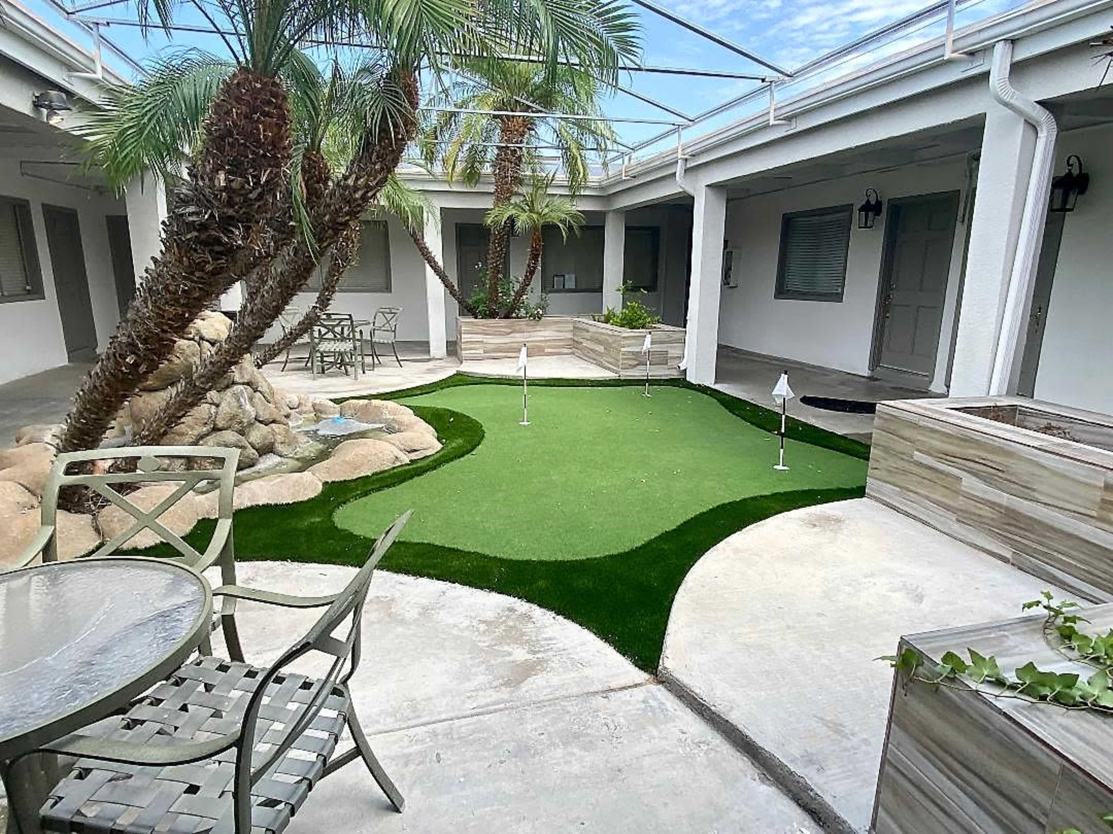 The image shows a landscaped courtyard featuring a putting green, potted palm trees, and seating areas with a table and chairs.