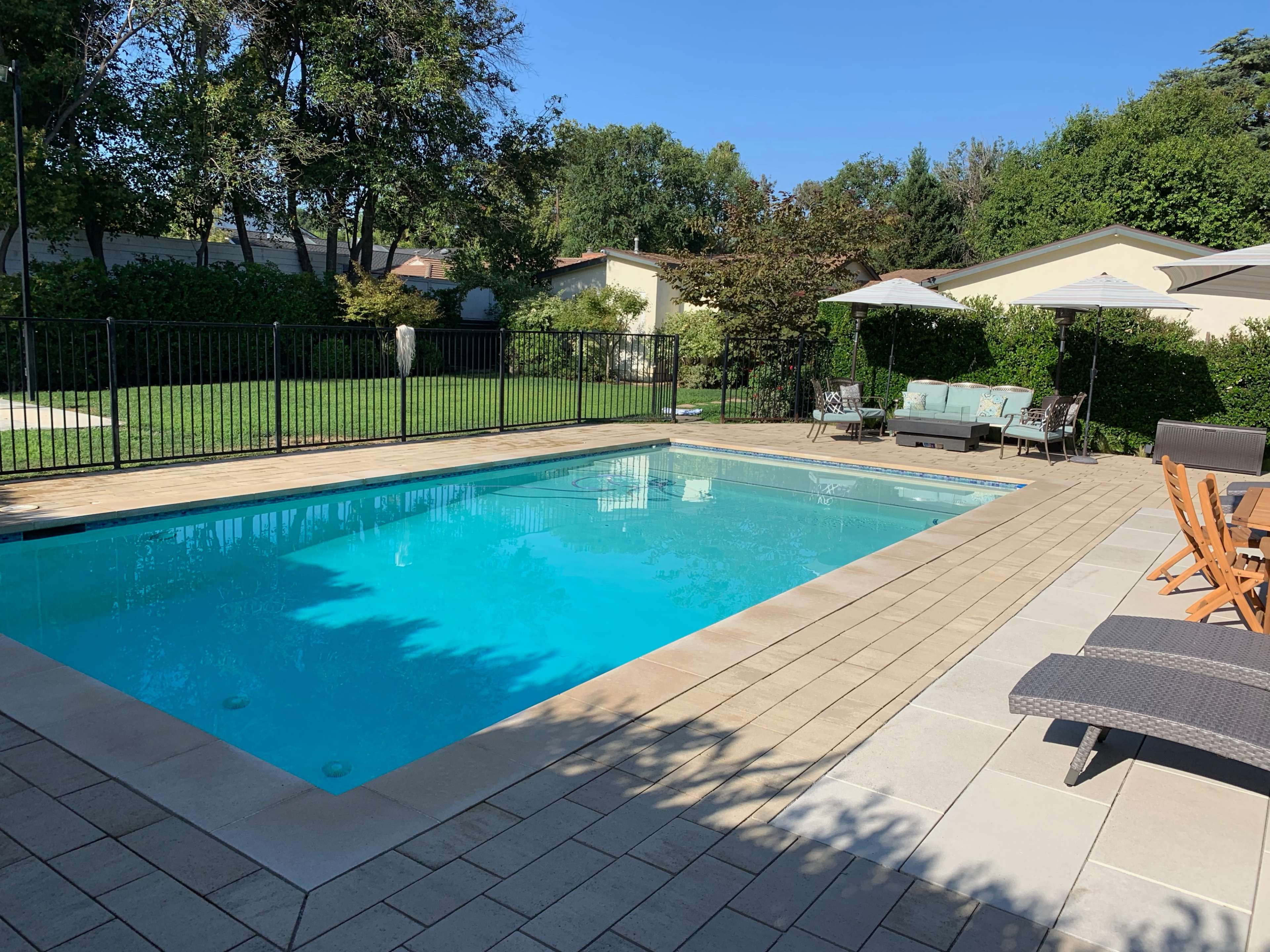 A rectangular swimming pool is surrounded by a stone patio, with lounge chairs and outdoor seating near a green lawn and hedges.