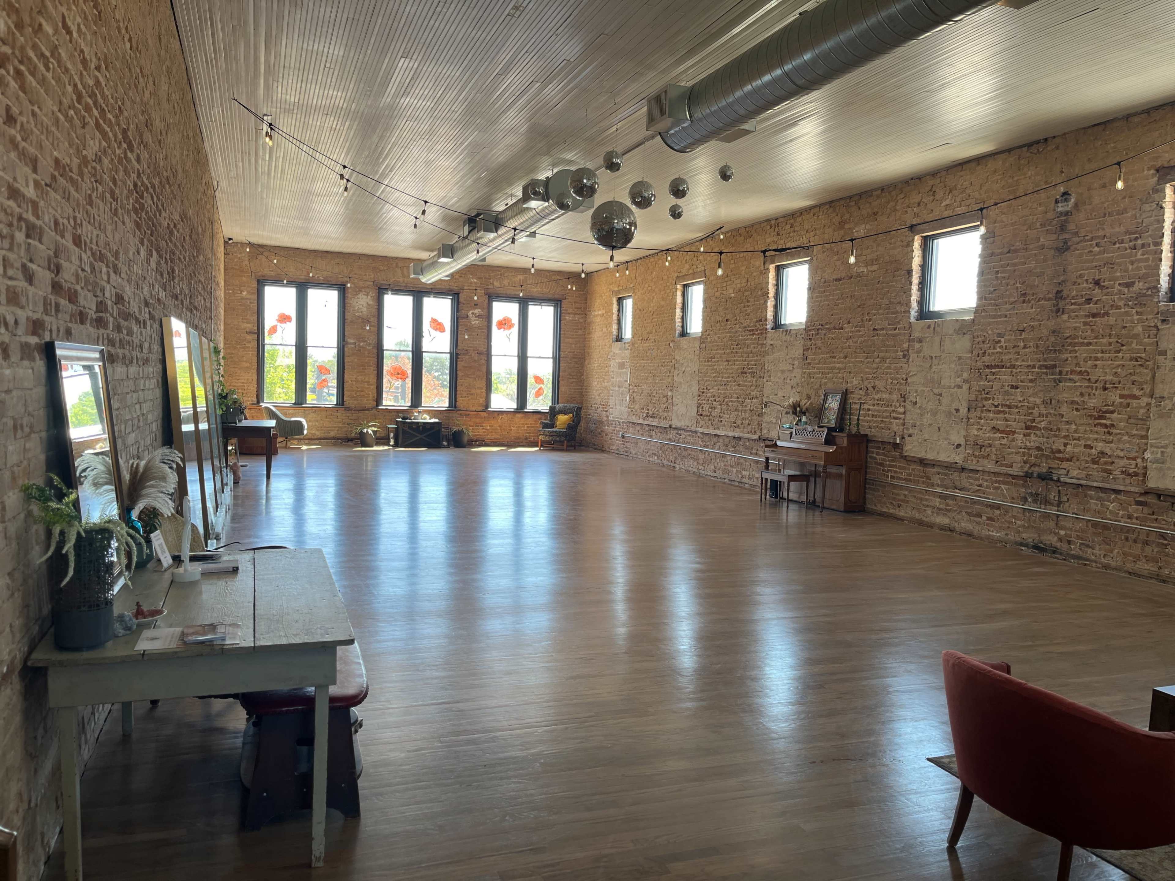 The image shows a spacious, airy room with wooden floors, exposed brick walls, large windows, and decorative mirrors.