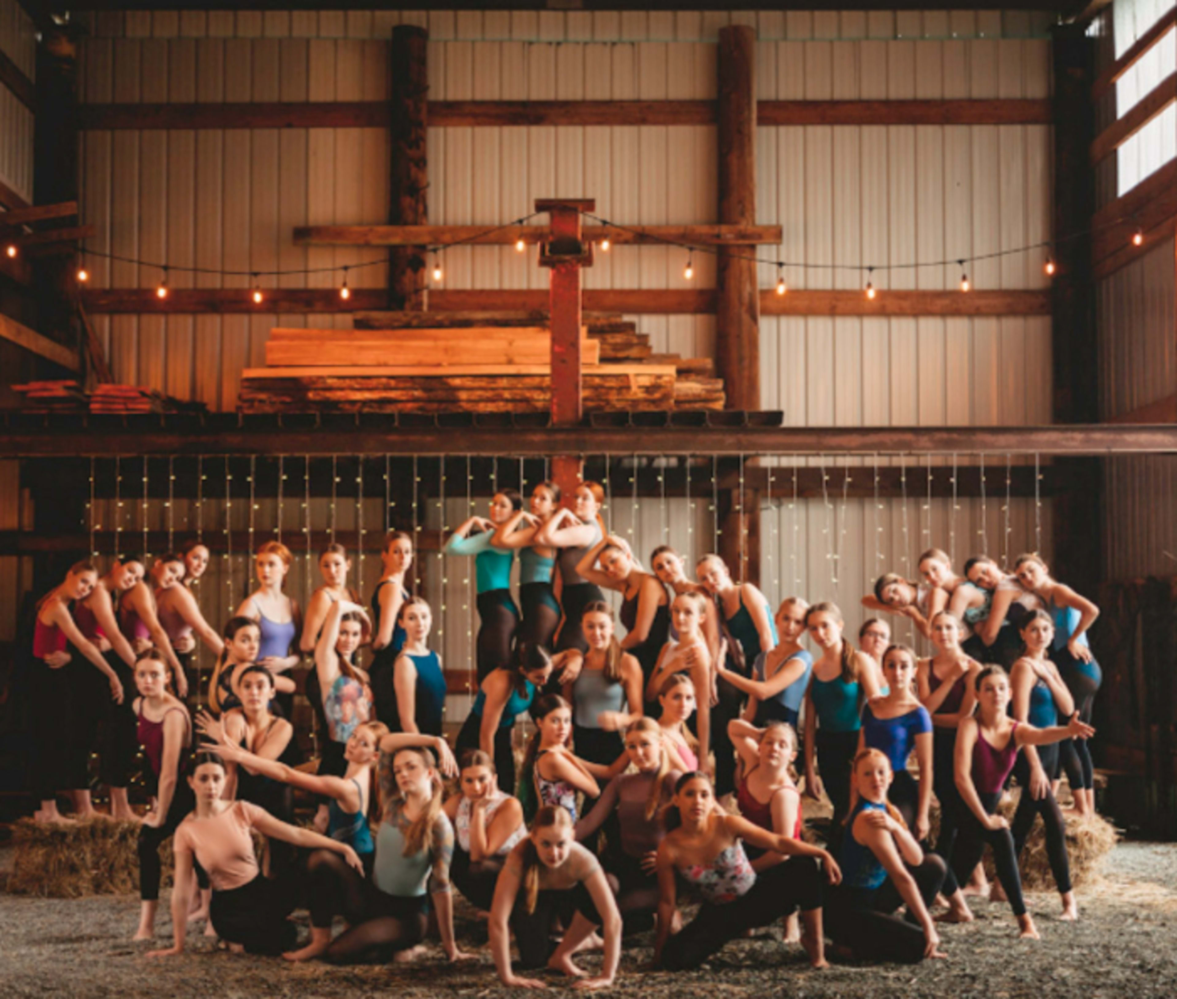 A large group of dancers poses together in a rustic warehouse featuring wooden beams and string lights.