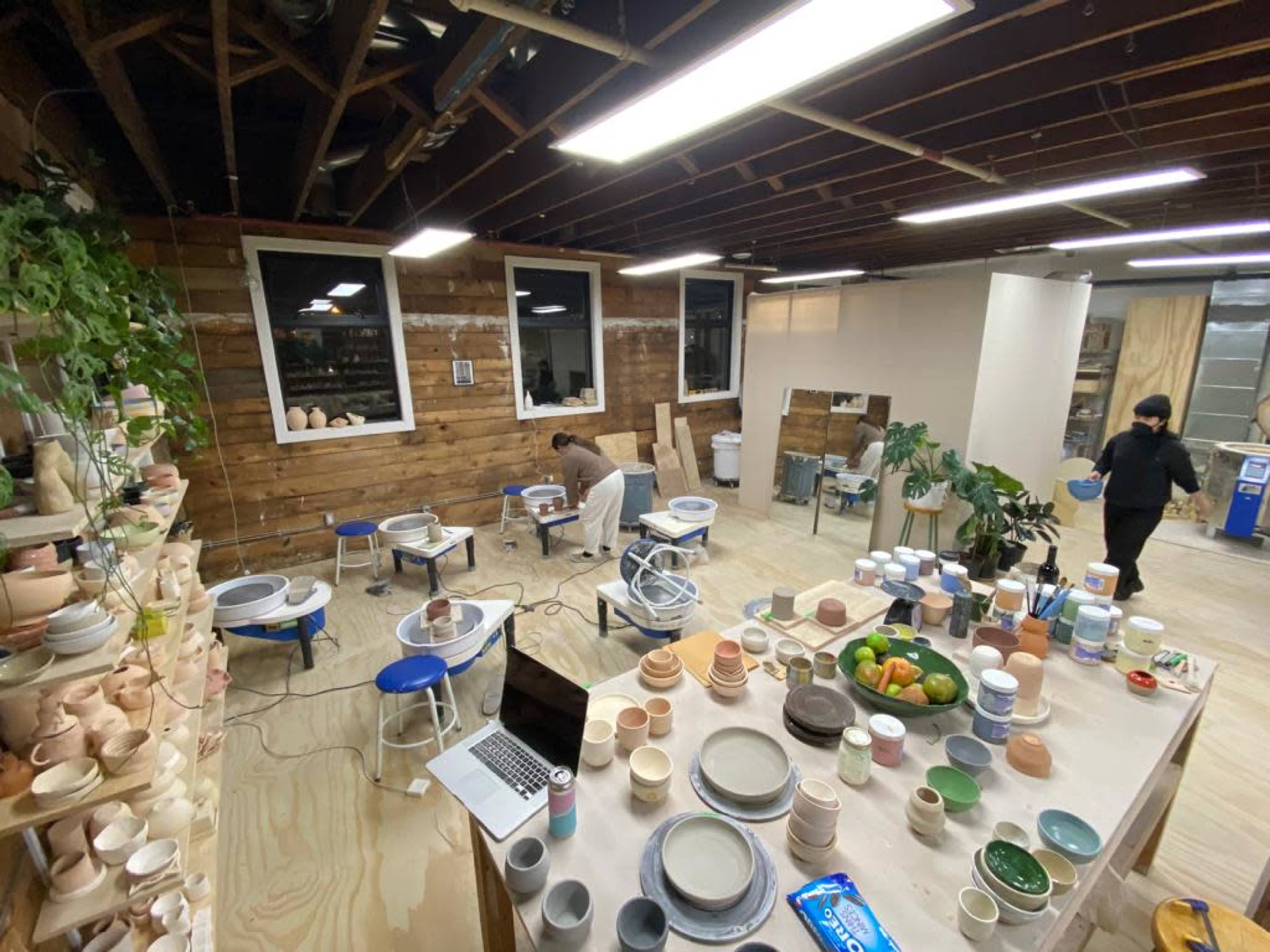 A pottery workshop with several people working at potter's wheels and various ceramic items displayed on tables around the room.