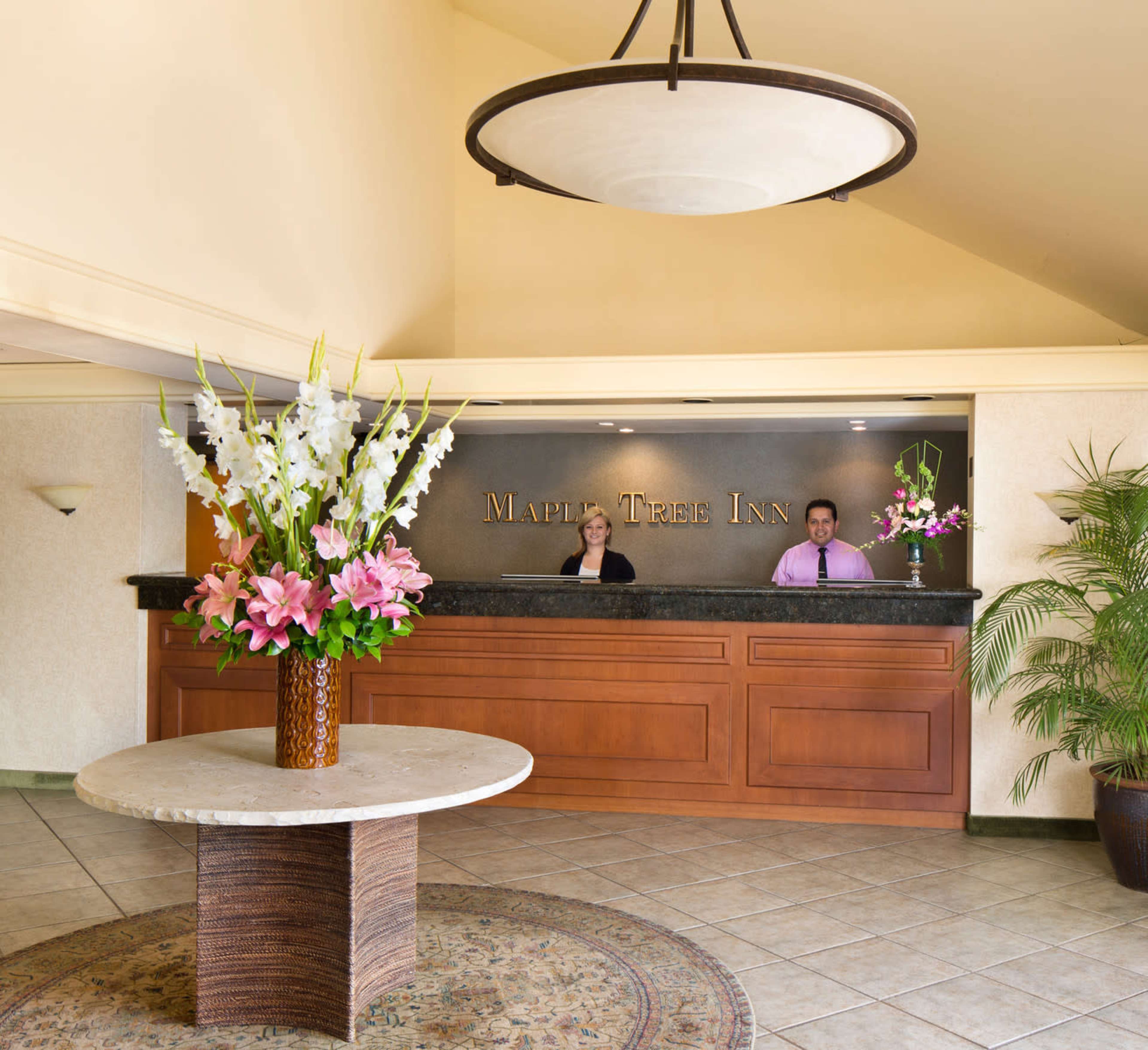The image shows a hotel reception area with a front desk, two staff members, and a large flower arrangement on a round table in front of the desk.