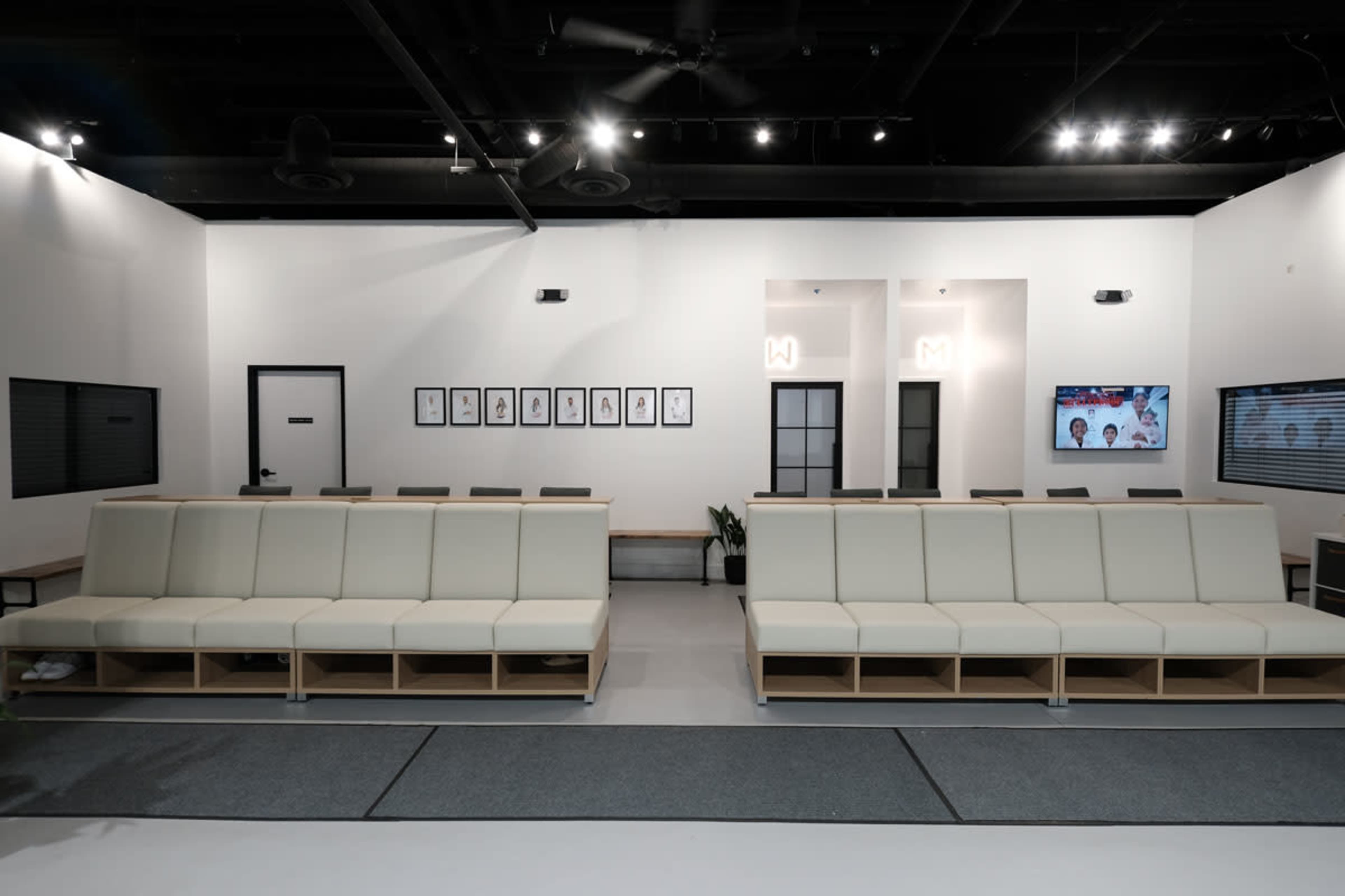 The image shows a modern waiting area with large, white benches, framed artwork on the walls, and a television mounted in a well-lit space.