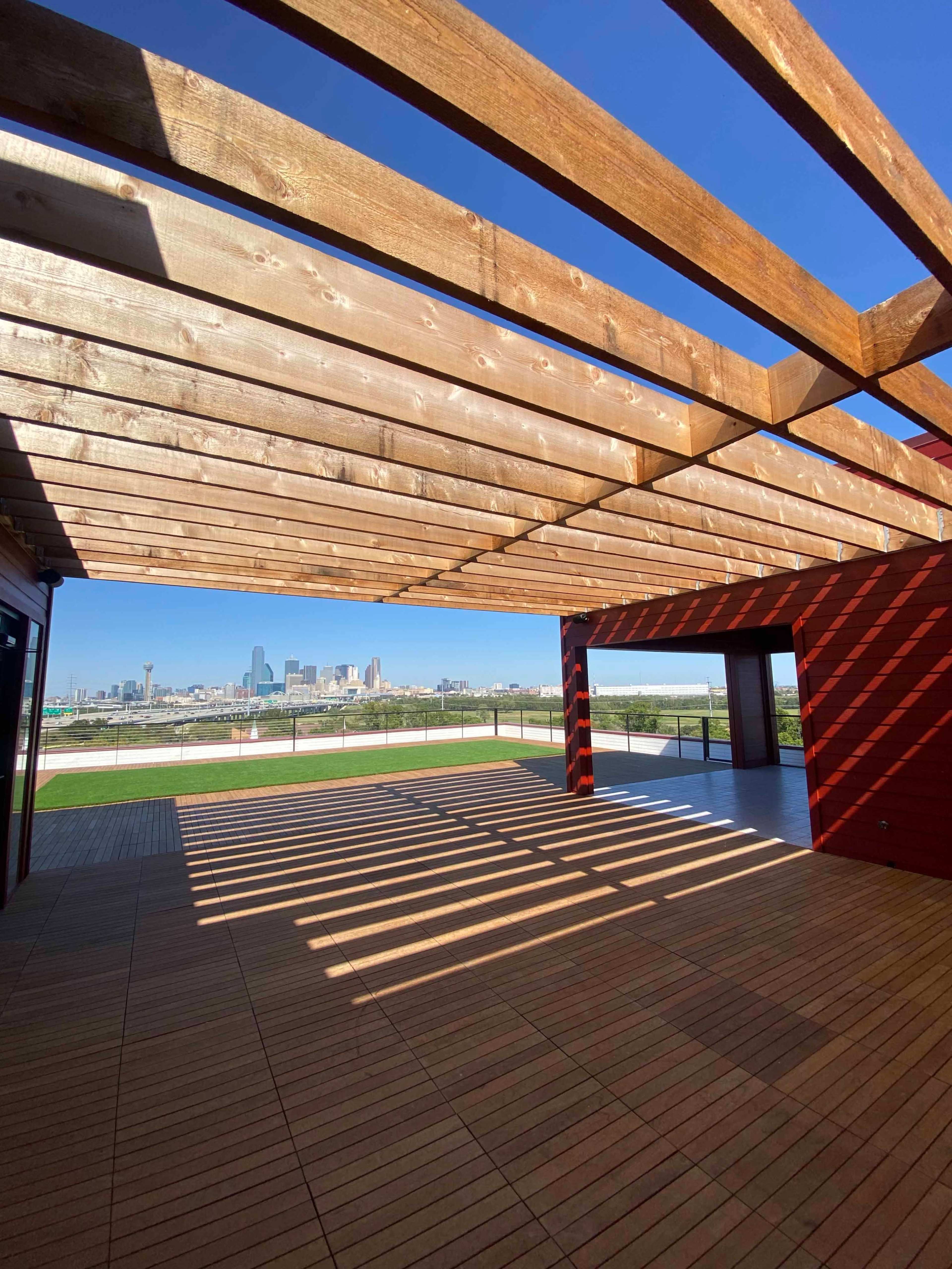 An open wooden structure with beams overhead, casting shadows onto a spacious deck that opens to a view of a city skyline in the background.