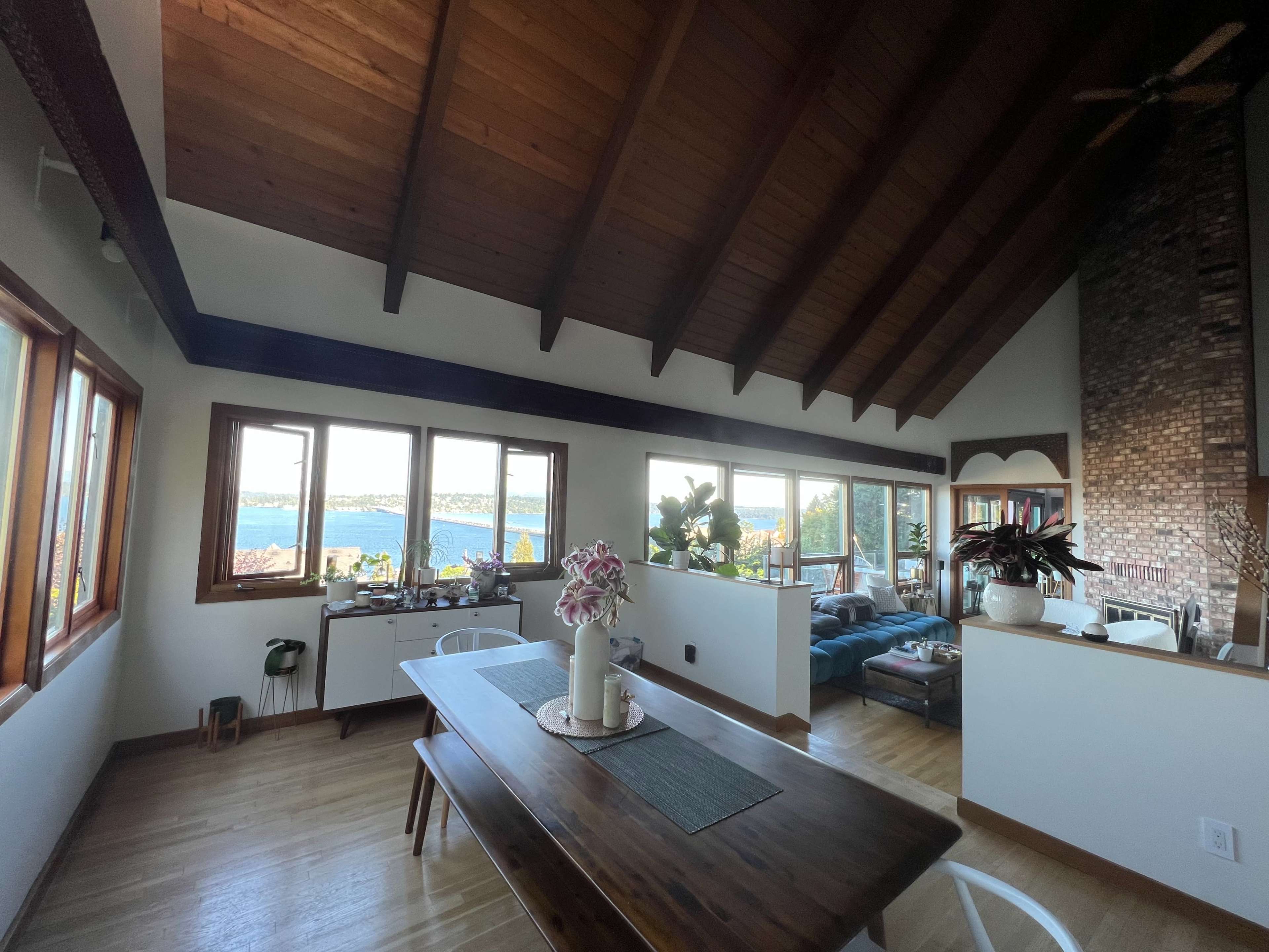 A cozy living space features a wooden dining table and a sectional sofa arranged near large windows overlooking a body of water.