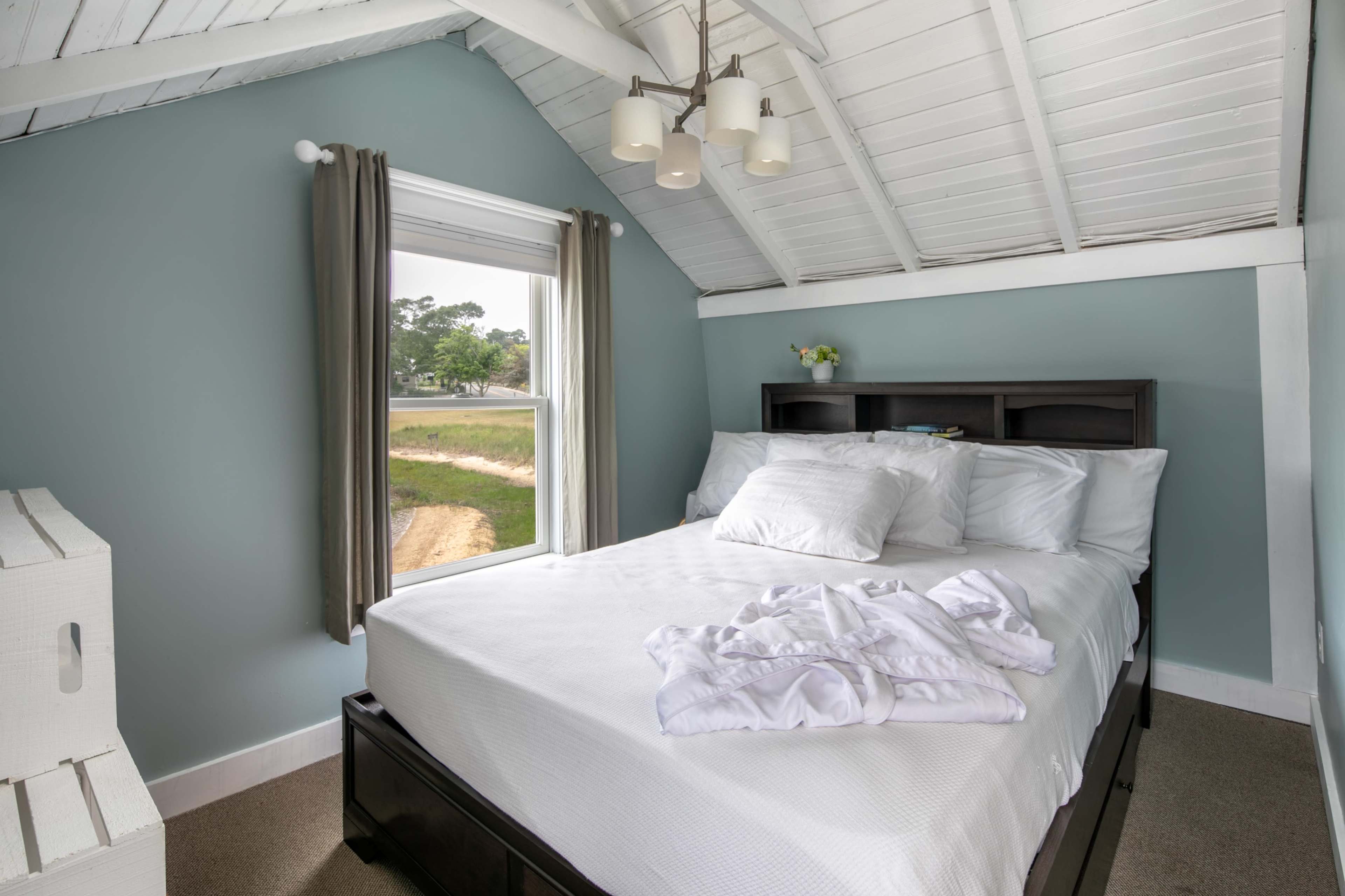 A neatly arranged bedroom features a bed with white linens and a large window overlooking a grassy area.