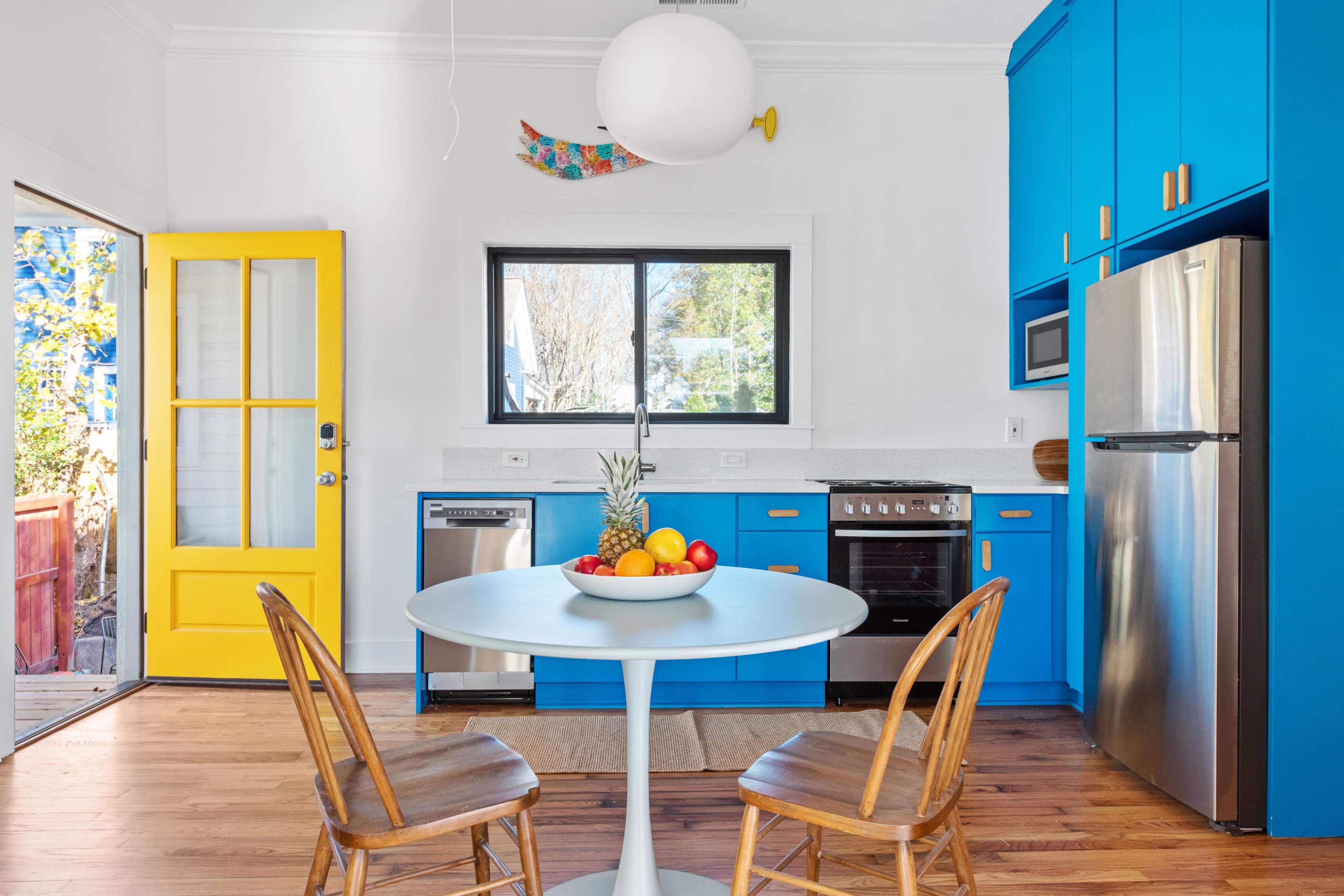 A bright kitchen features blue cabinets, stainless steel appliances, a white round table with a fruit bowl, and a yellow door leading to an outdoor area.