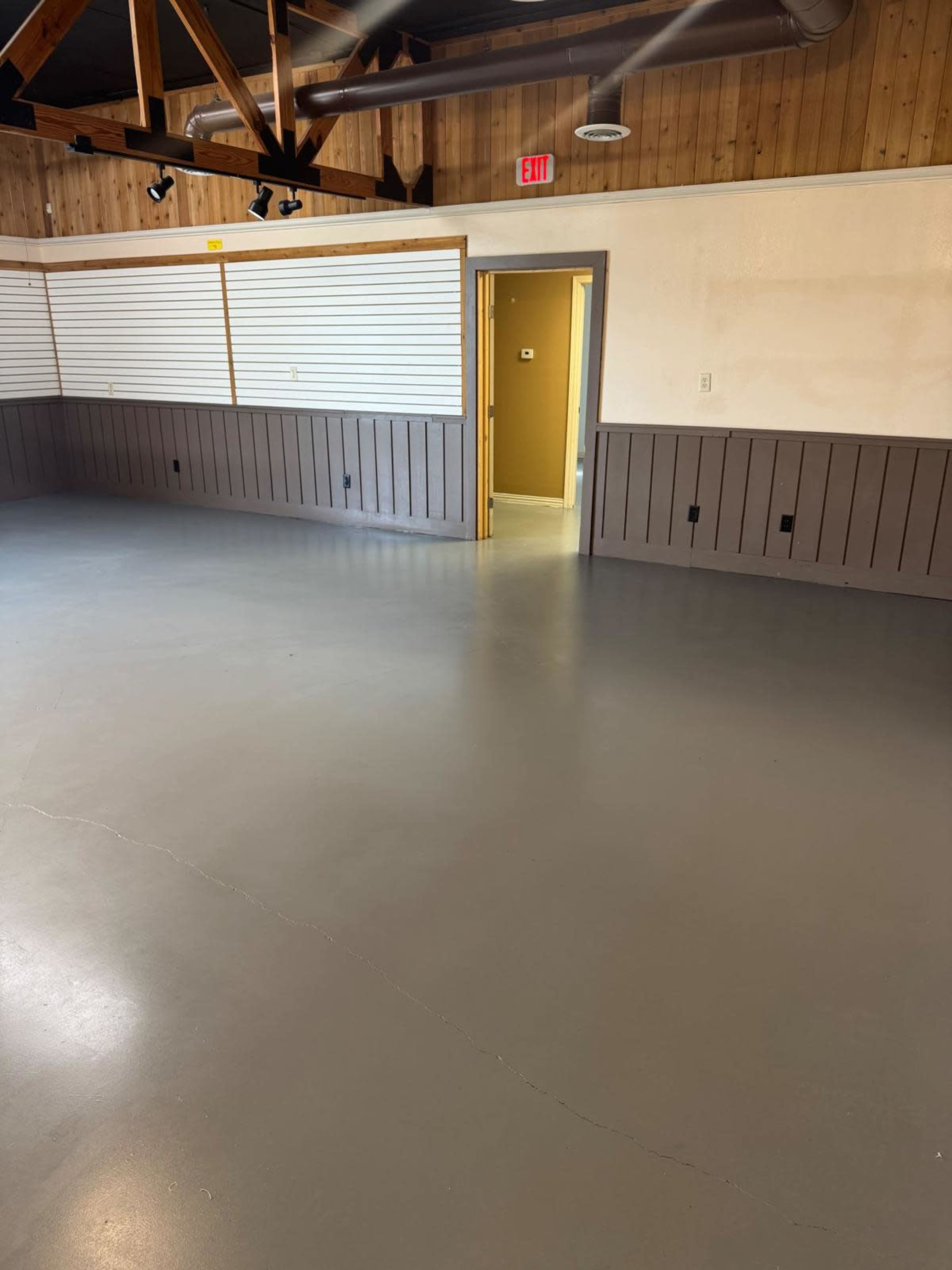 The image shows an empty room with plain gray flooring, wooden paneling on the lower half of the walls, and a door leading to another room in the background.