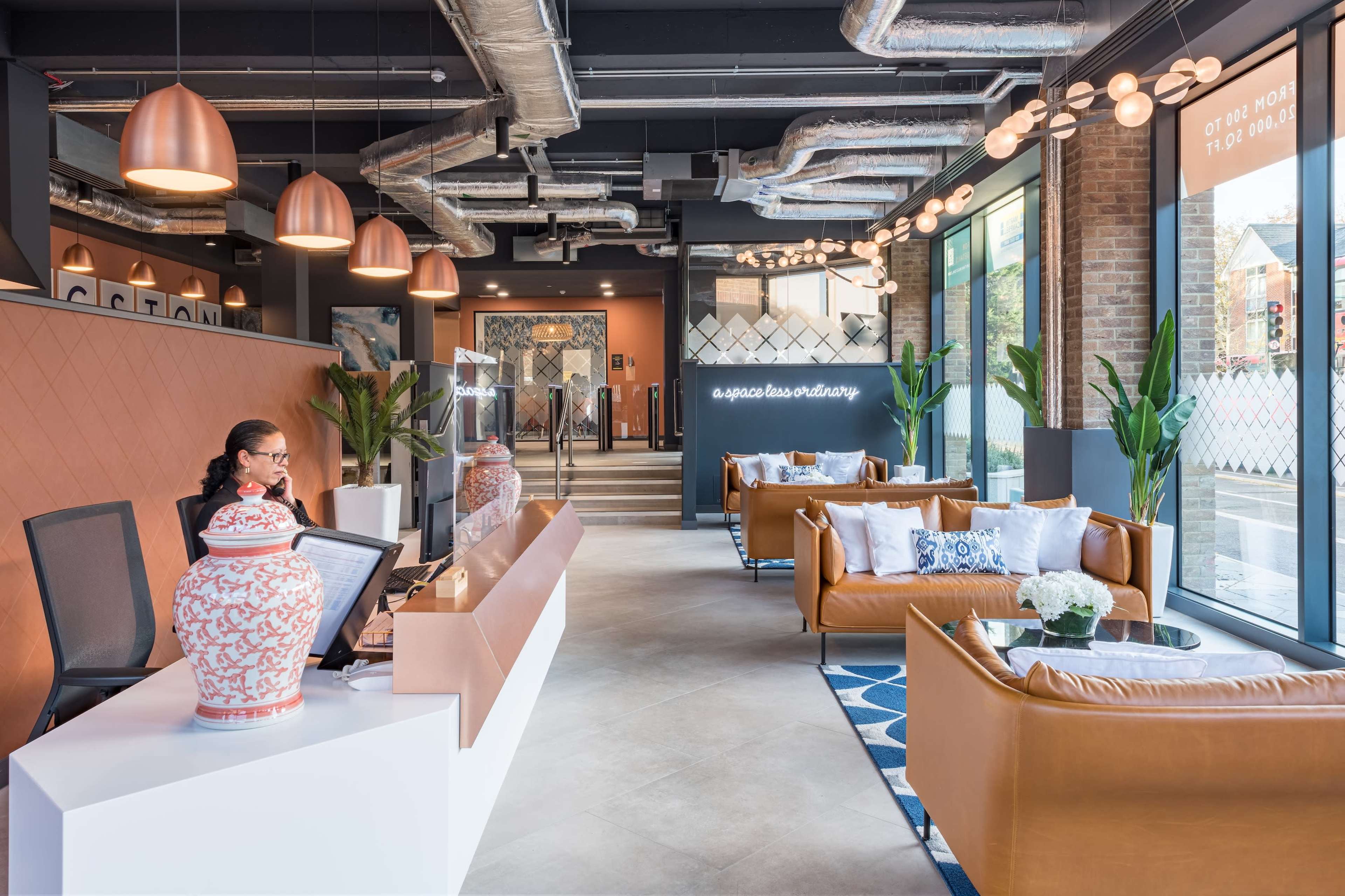 A modern reception area with a desk, seating arrangements with brown leather sofas, decorative plants, and large windows allowing natural light.