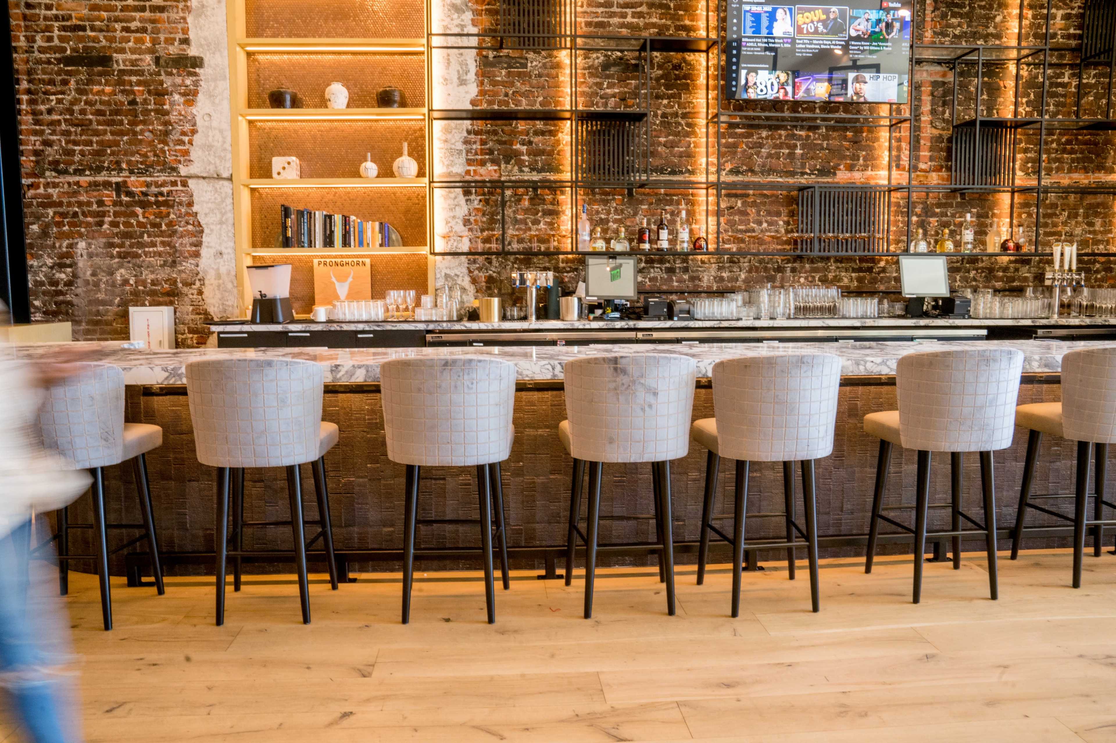 A modern bar with high-backed stools lined up along a counter, set against a brick wall with shelves displaying decorative items and a television screen above.