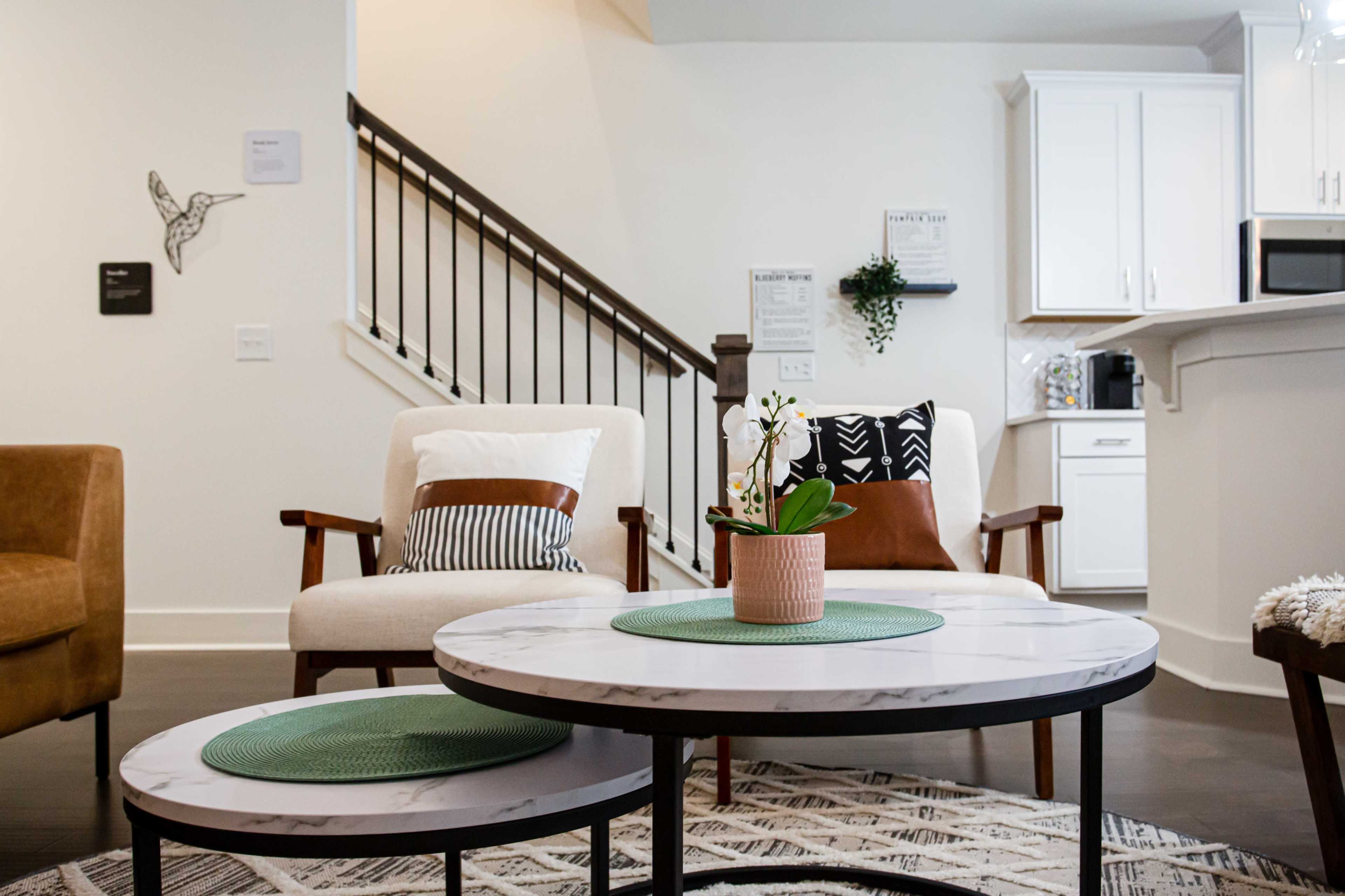 A modern living room featuring two armchairs, a round coffee table with green placemats, and a kitchen area in the background.