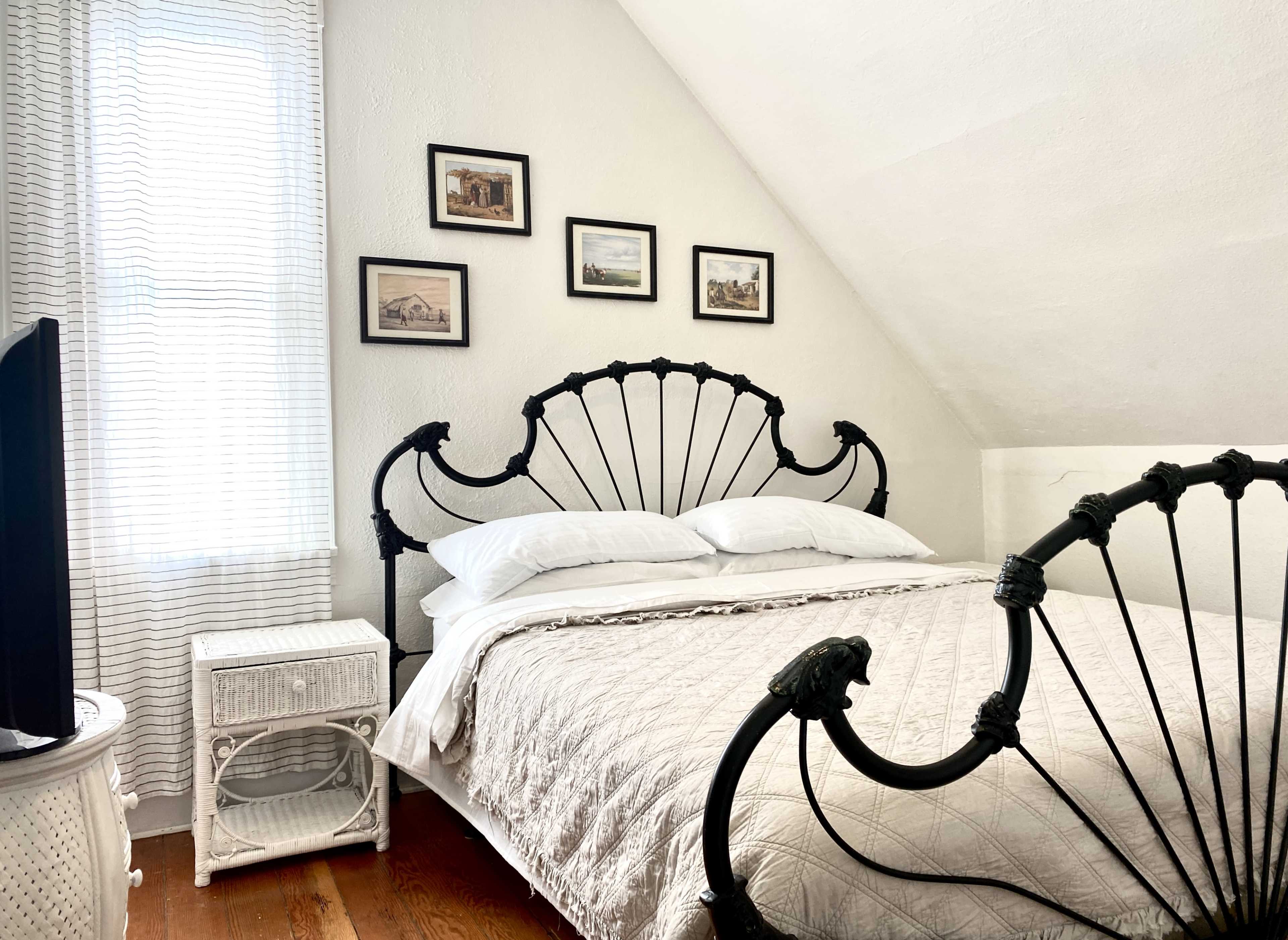 A bedroom features a black iron bed frame with white bedding, accompanied by a nightstand and framed pictures on the wall.