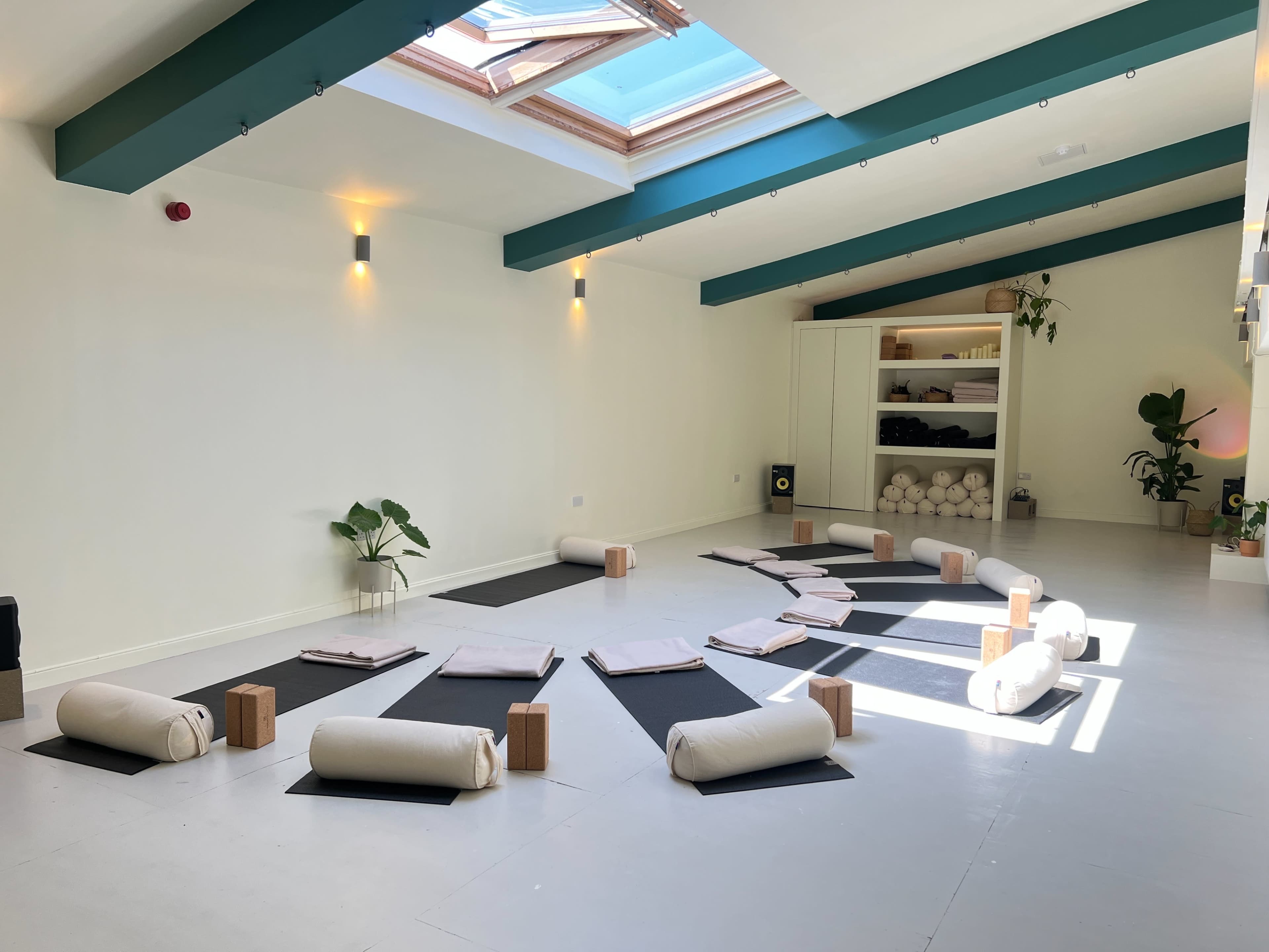 The image shows a spacious yoga studio with neatly arranged mats, bolsters, and props, illuminated by natural light from a skylight.